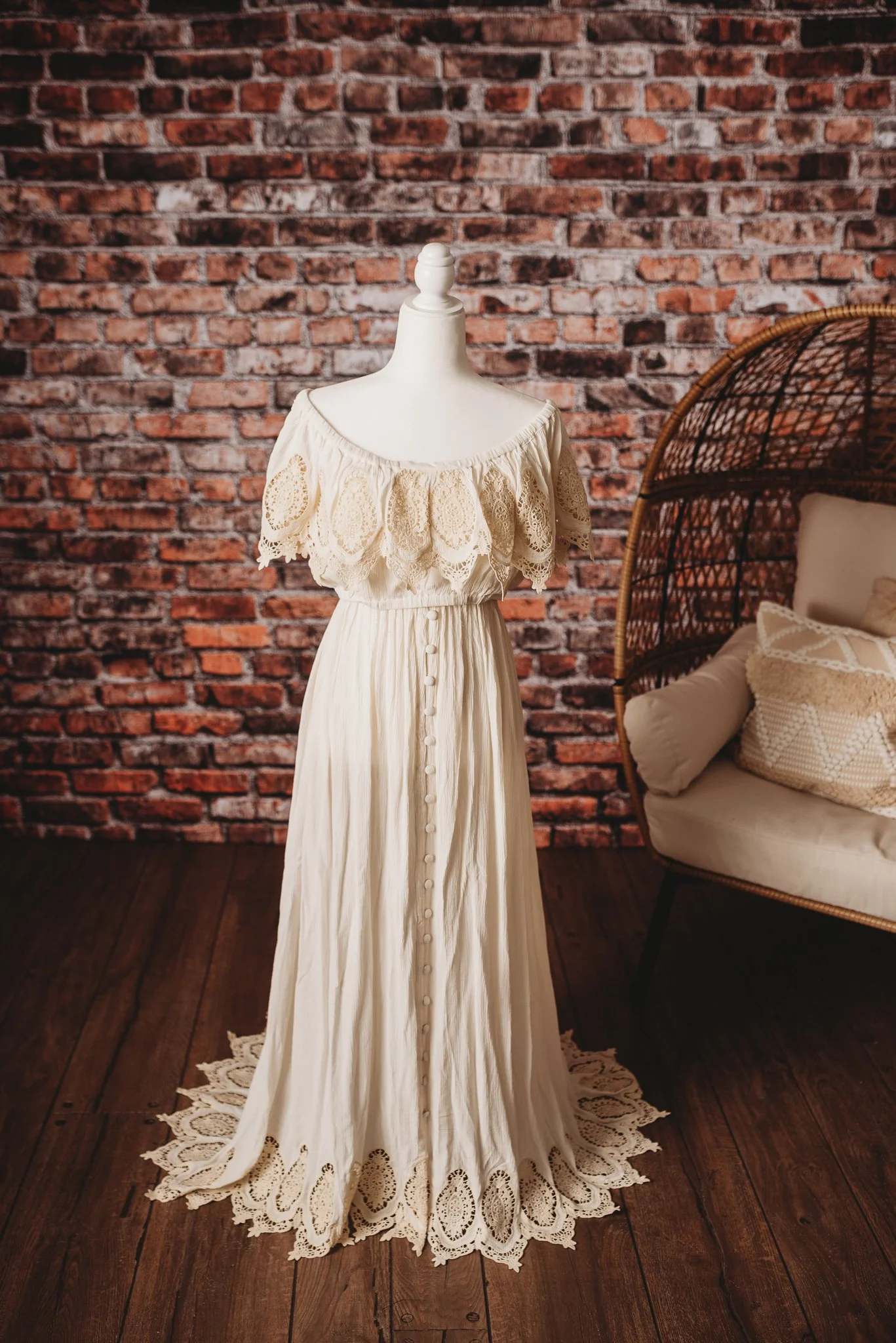 Off-shoulder lace maxi dress on mannequin in rustic setting with brick wall and wicker chair.