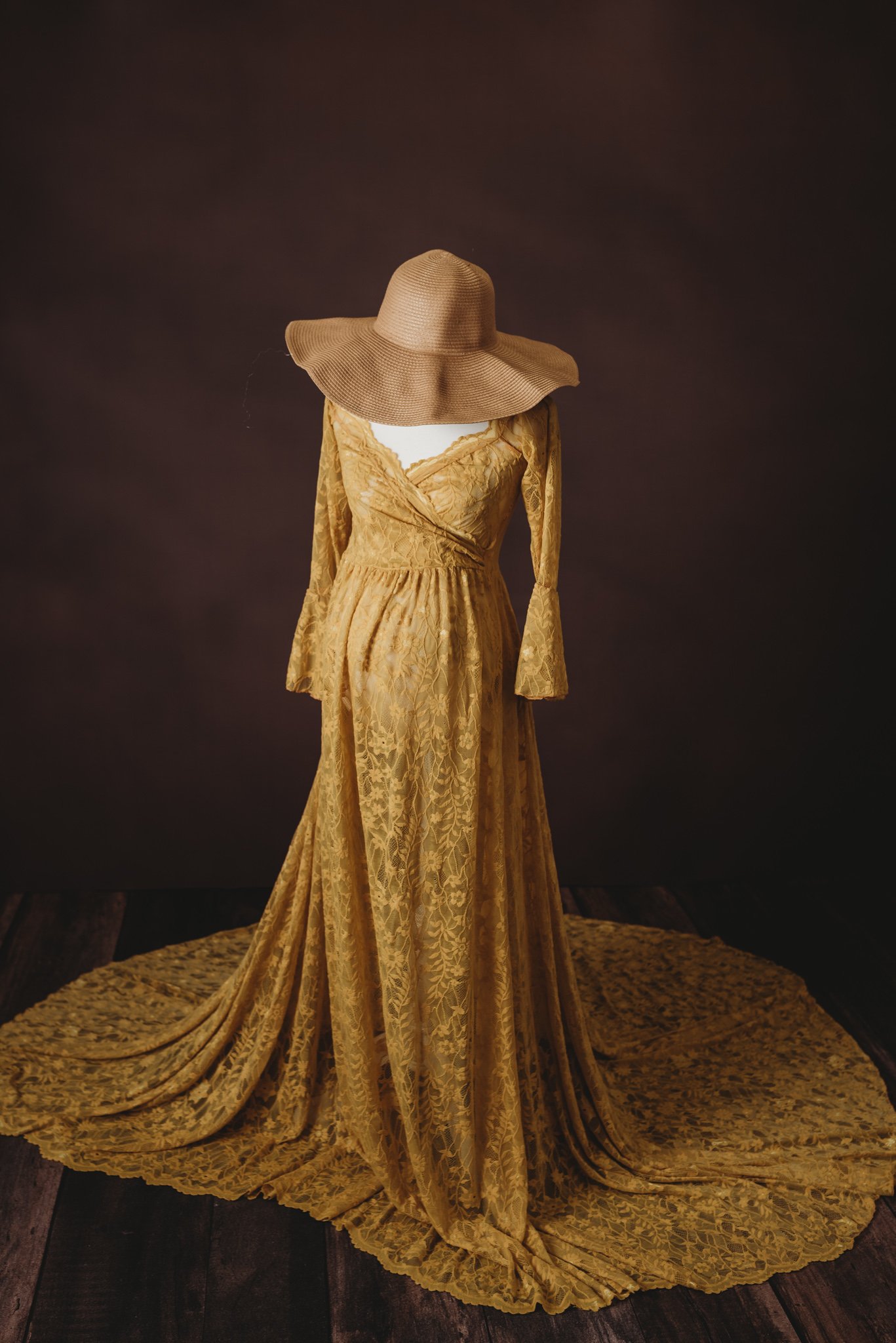 Elegant mustard-yellow lace maternity gown with a wide-brimmed hat on a dark background.