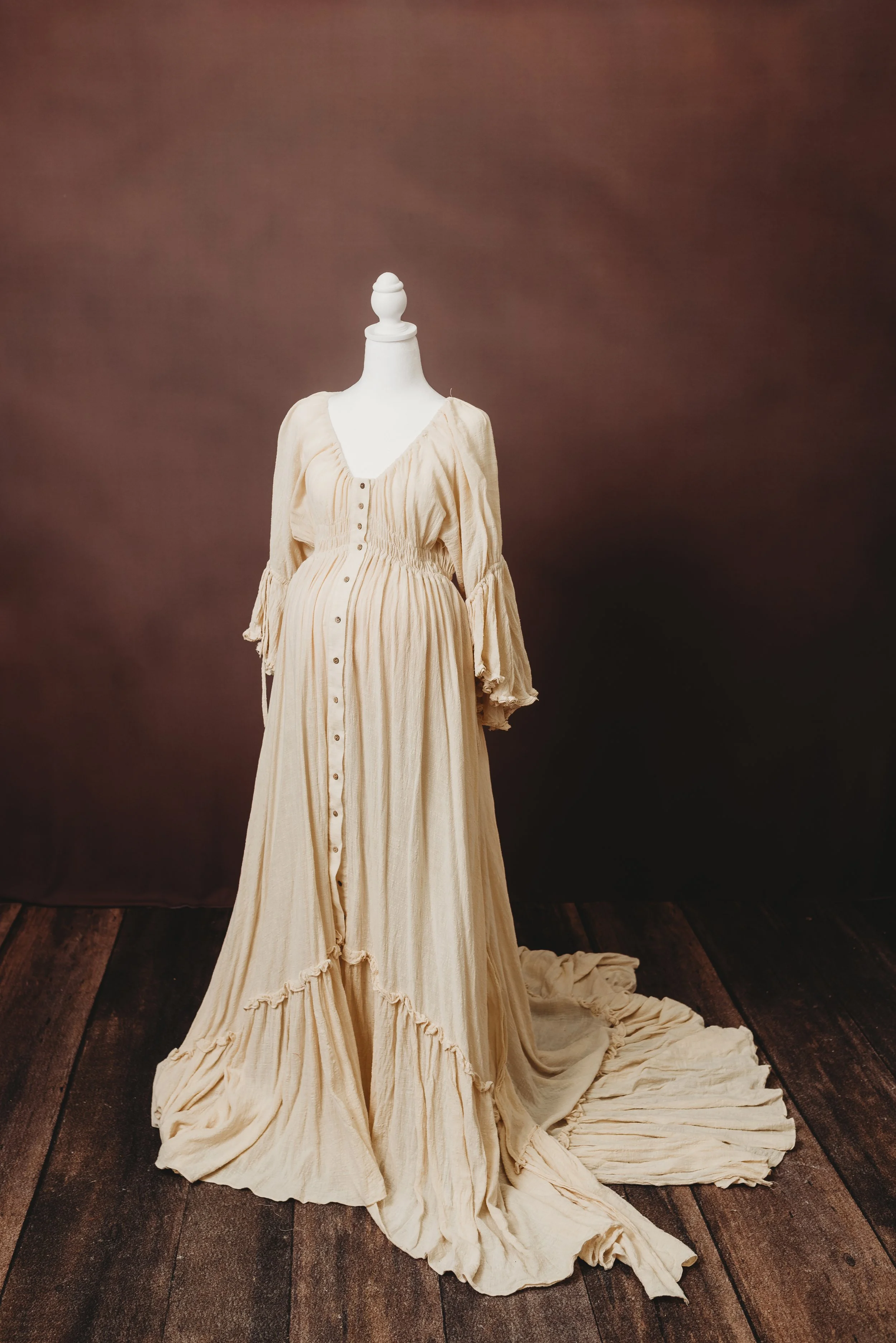 A long beige vintage dress with buttons down the front displayed on a white mannequin, set against a brown backdrop on a wooden floor.