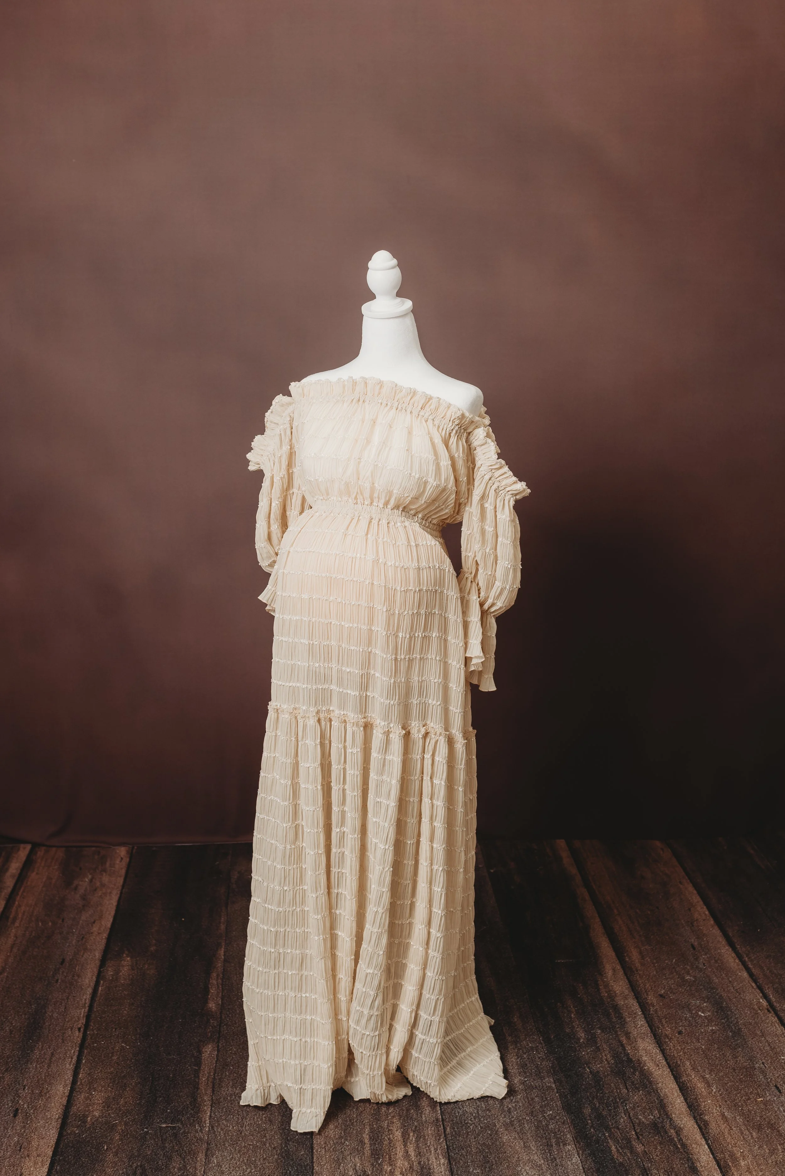 A mannequin wearing a long, cream-colored, pleated dress with ruffled details and an off-the-shoulder neckline, set against a brown backdrop with a wooden floor.