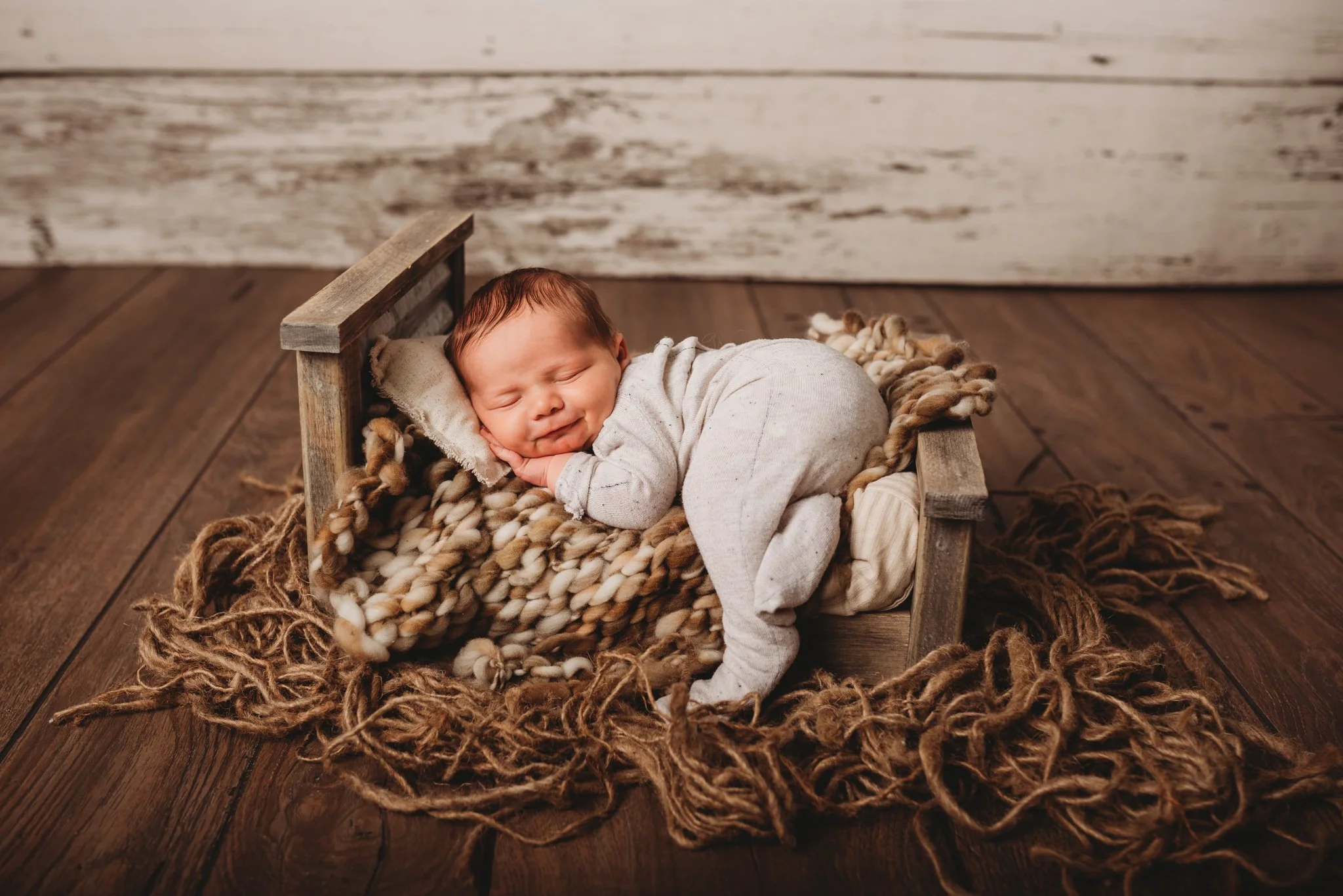 Sleeping newborn in a small wooden bed, wrapped in a knitted blanket on wooden flooring.