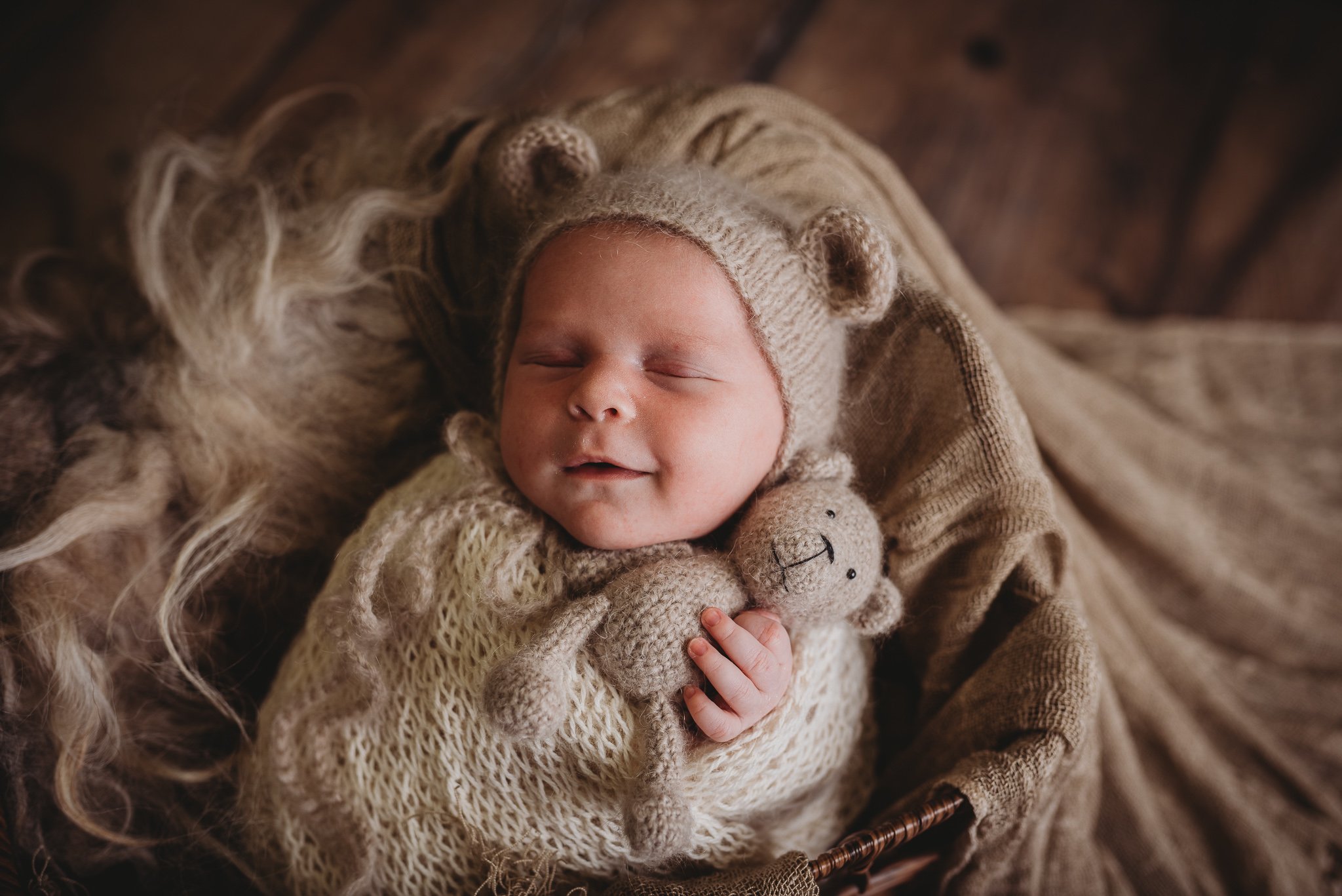 Sleeping baby in knitted bear hat, holding a small stuffed bear toy, wrapped in a cream blanket.