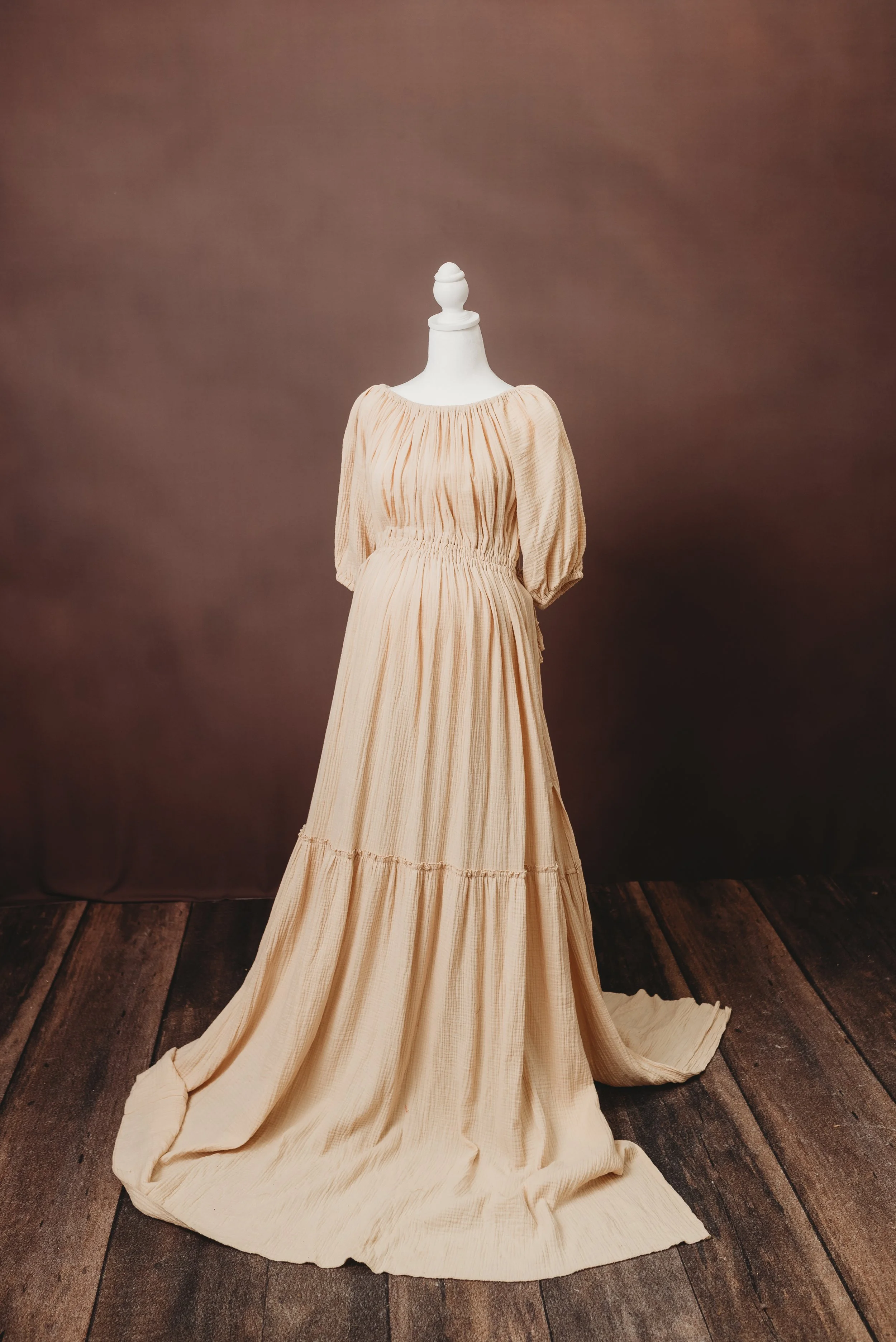 A vintage-style, beige, pleated dress displayed on a mannequin against a plain, dark background. The dress has a long, flowing skirt that pools on a wooden floor.