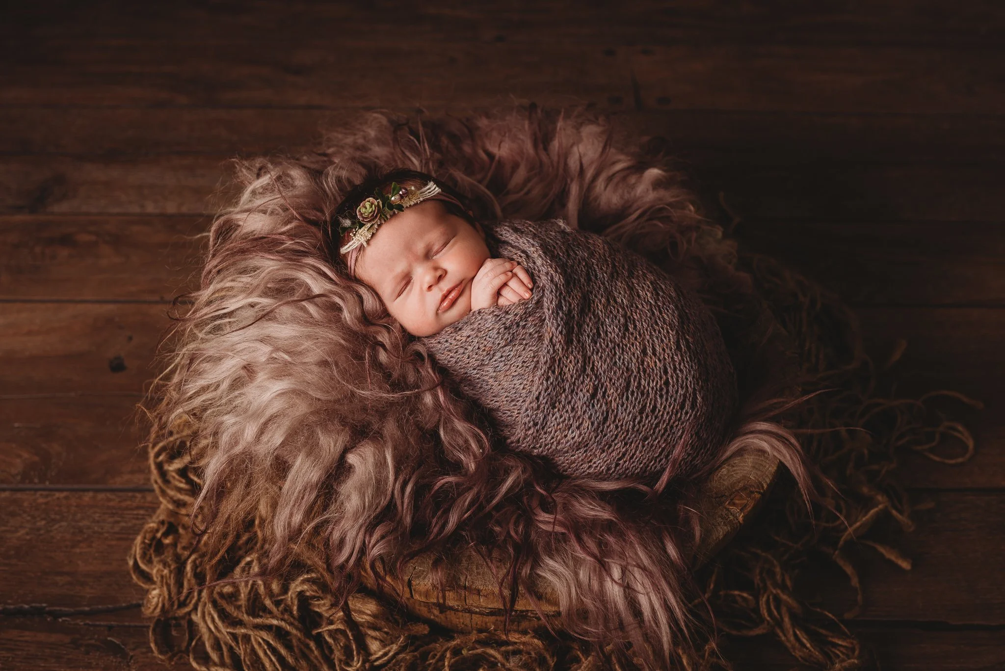 Sleeping baby wrapped in blanket, adorned with floral headband, lying on furry blanket on wooden floor.