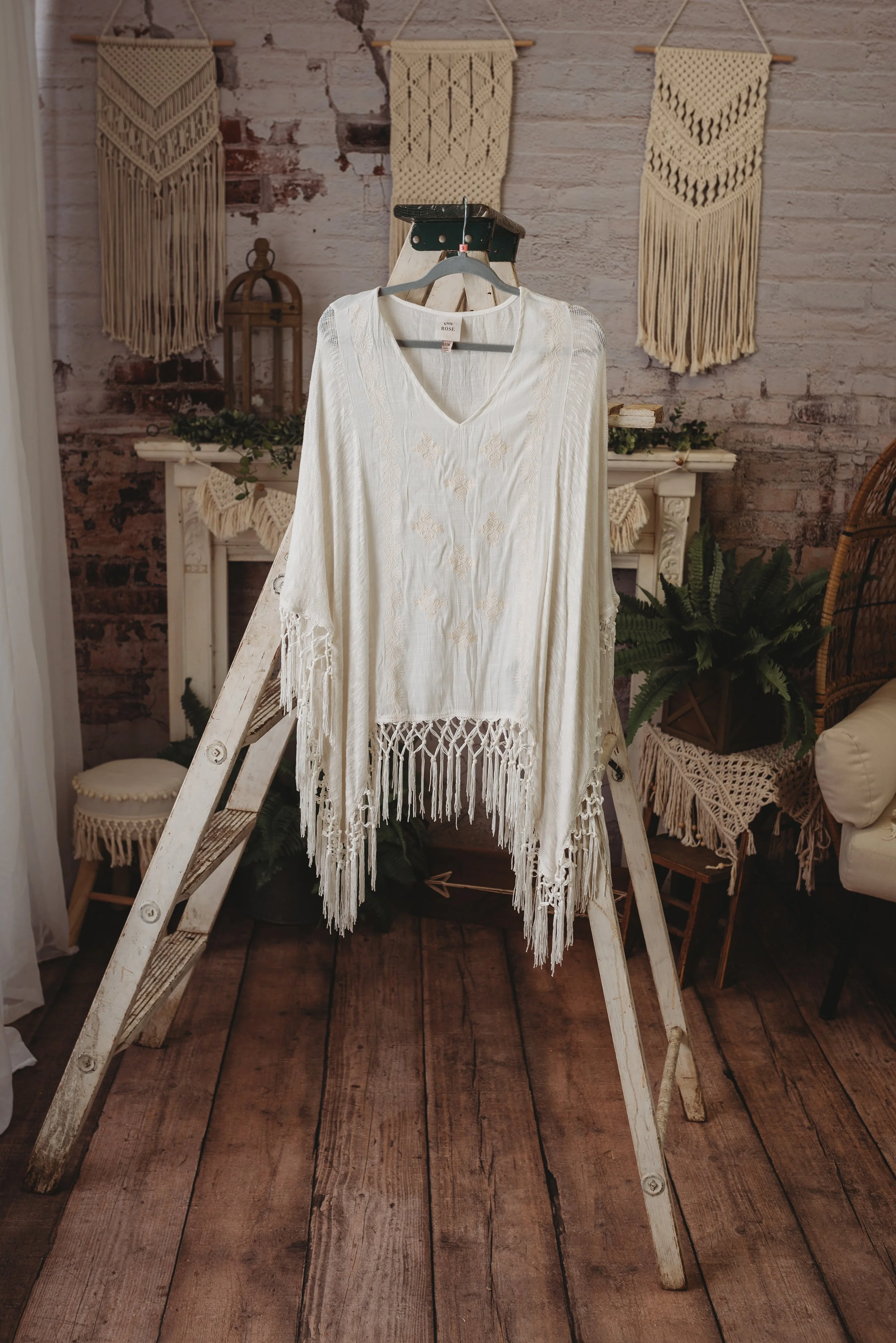 Bohemian-style white poncho with fringe hanging on a vintage wooden ladder in a rustic room with macramé wall hangings.