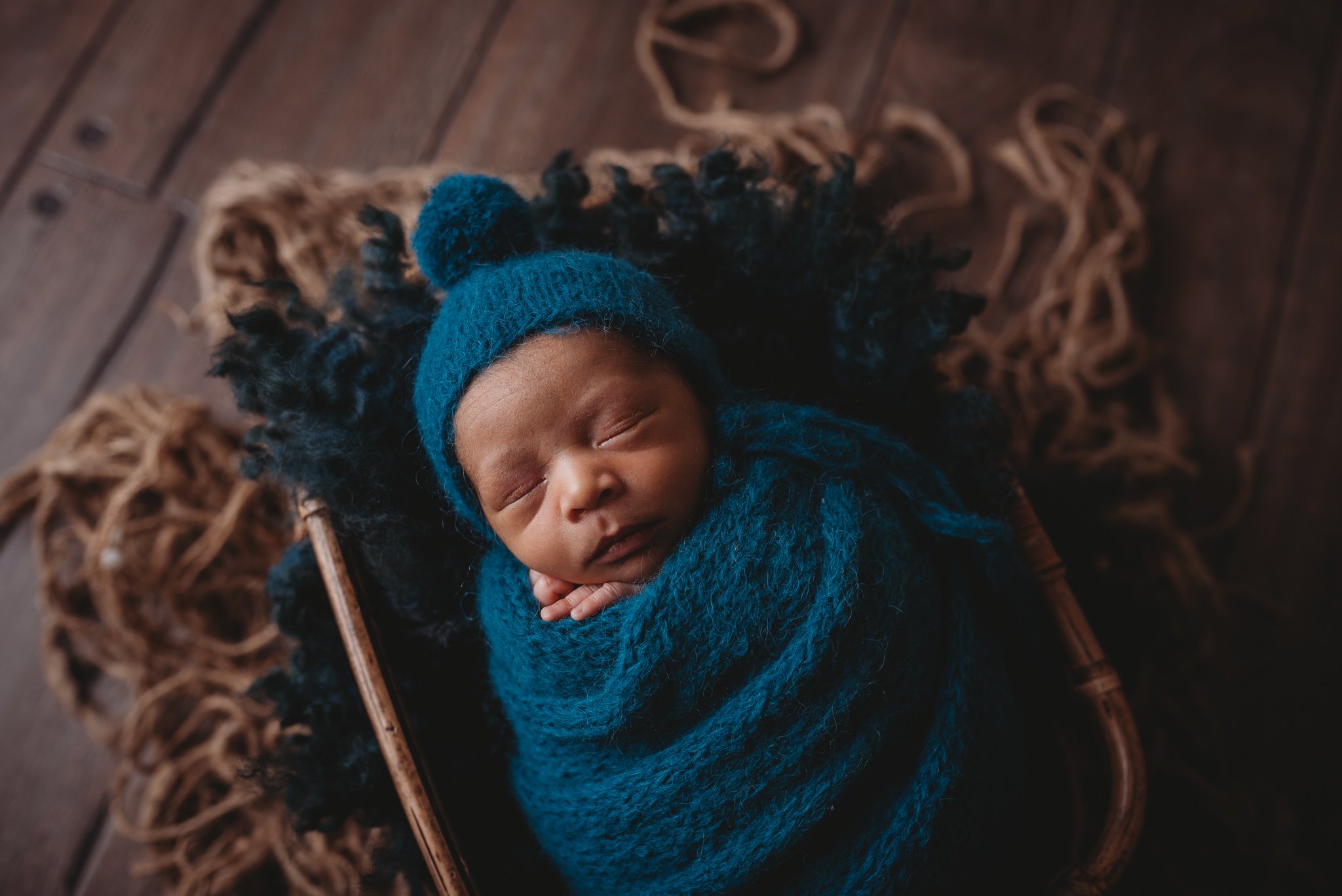 Newborn baby wrapped in blue blanket and hat, sleeping in a basket, with wooden floor background.