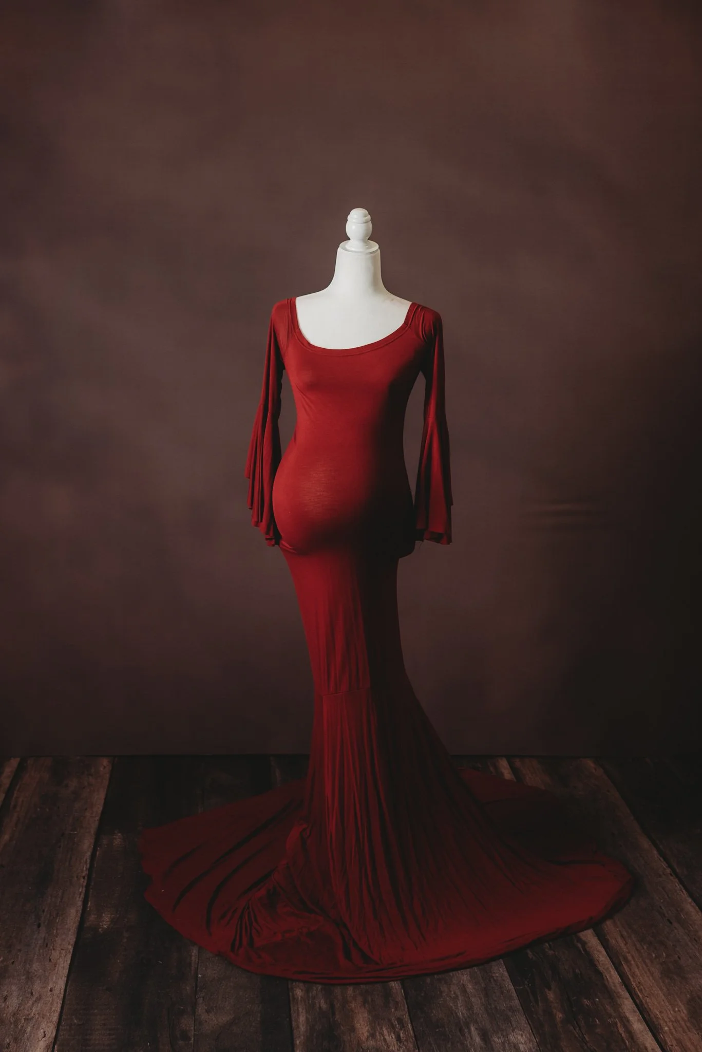Red maternity gown on mannequin, wooden floor, brown background.