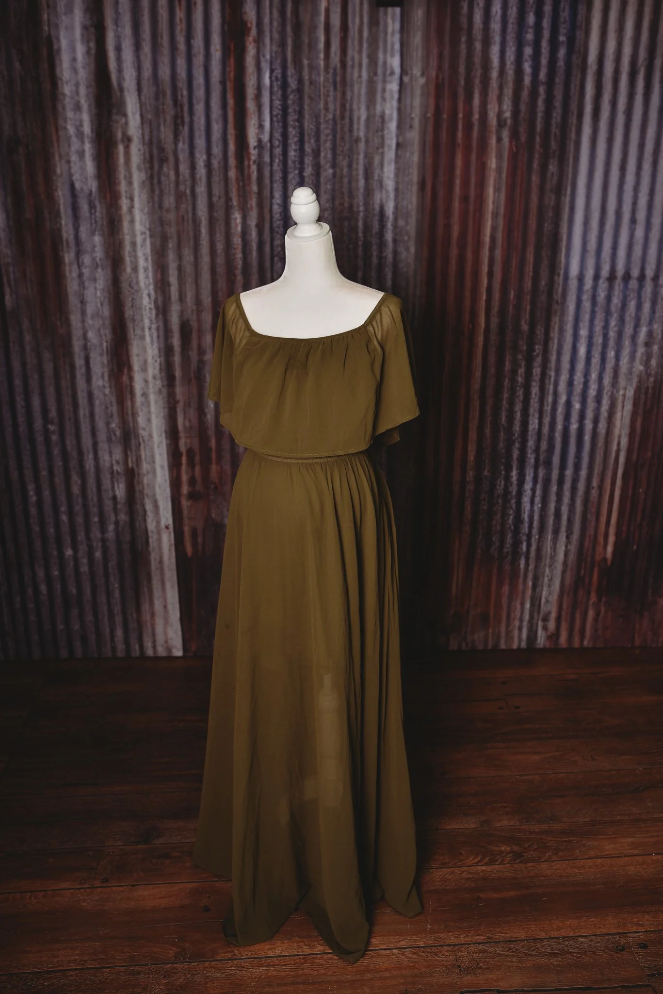 Mannequin displaying olive green dress with ruffled sleeves, wooden floor, corrugated metal background.