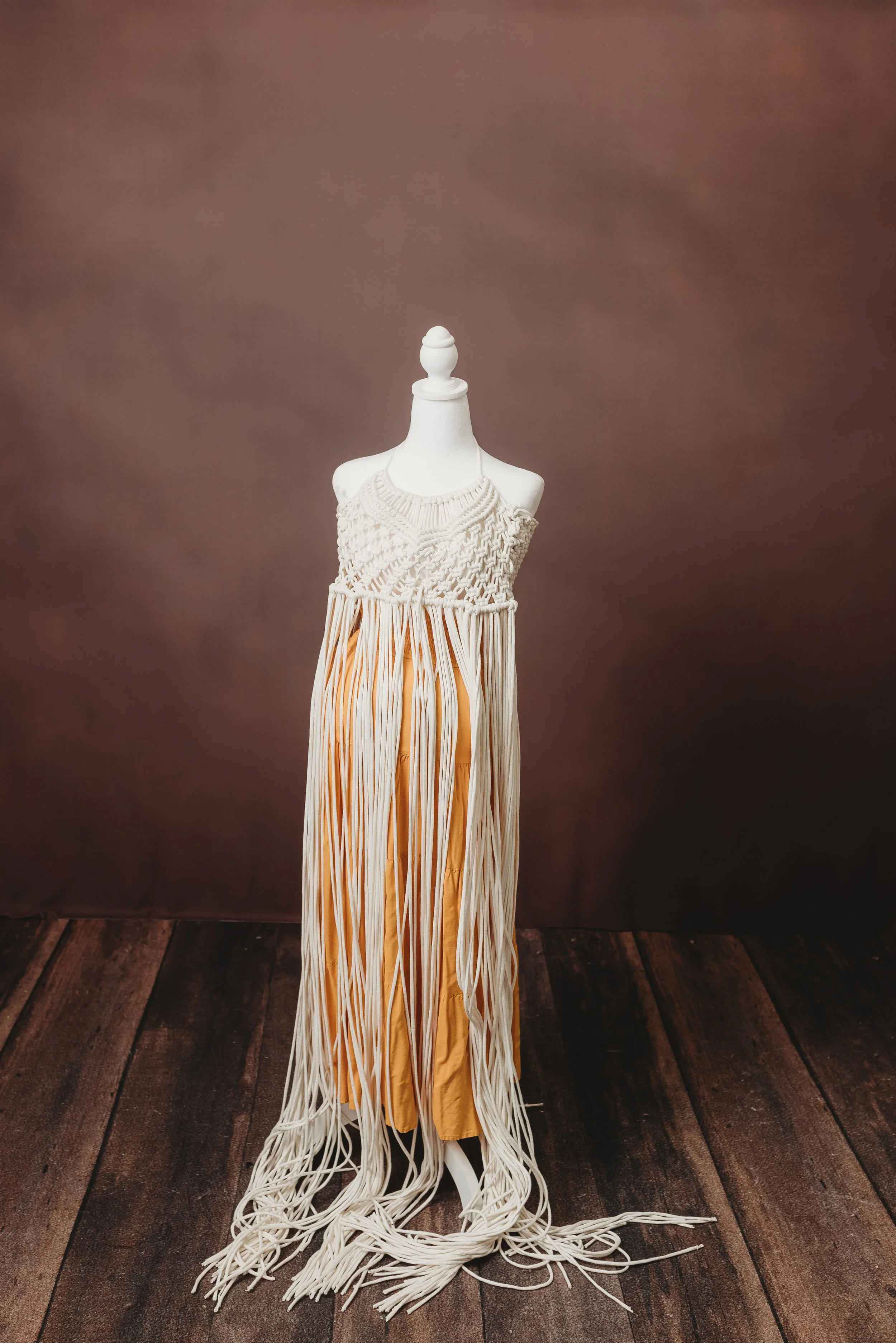 A mannequin draped with a layered, cream-colored knitted top and a long, flowing skirt with vertical stripes in yellow and white, set against a brown background on a wooden floor.