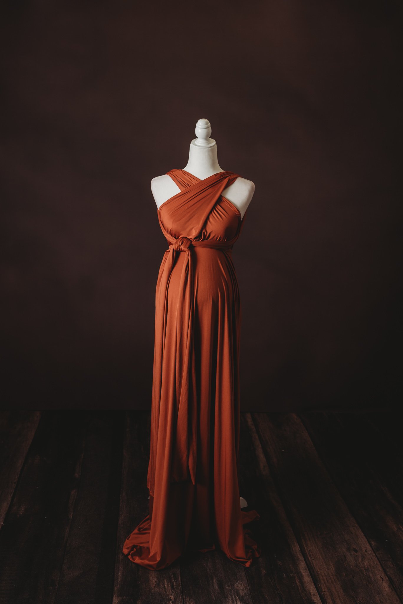 Mannequin wearing a rust-colored, long, wrap-style dress with cross-over straps, against a dark background.