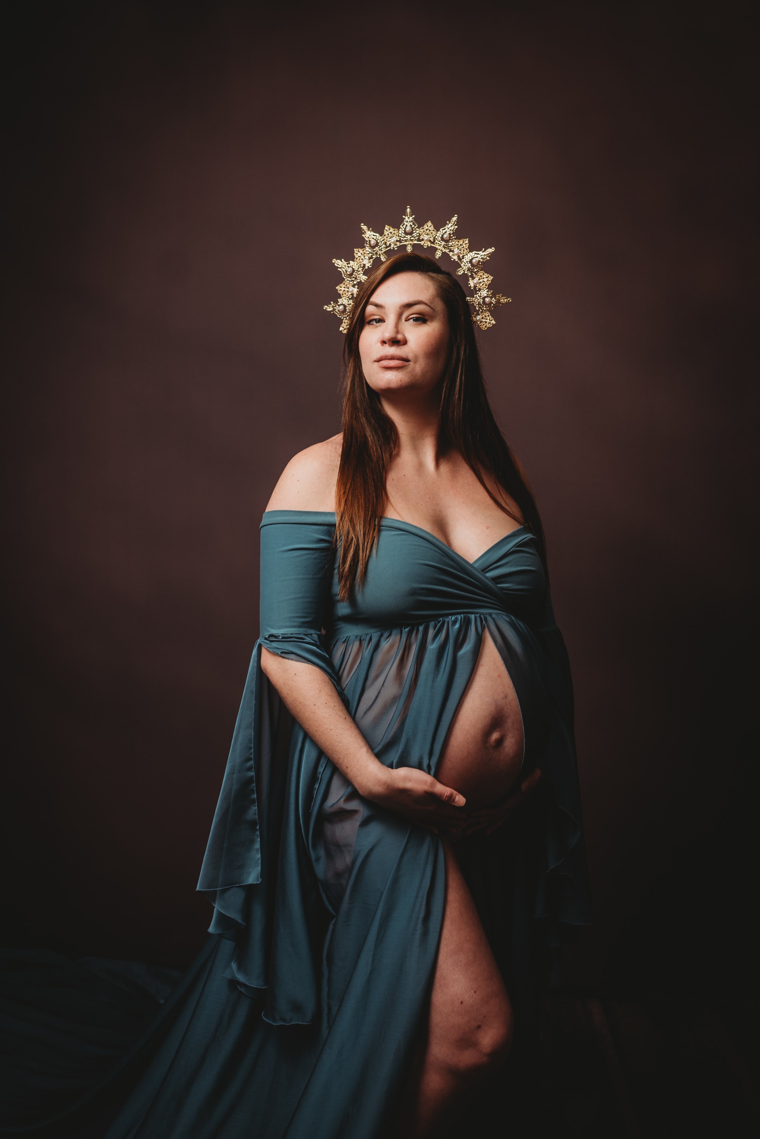 A woman with long brown hair wearing an off-the-shoulder blue dress and an ornate golden crown, holding her pregnant belly, standing against a dark, plain background.