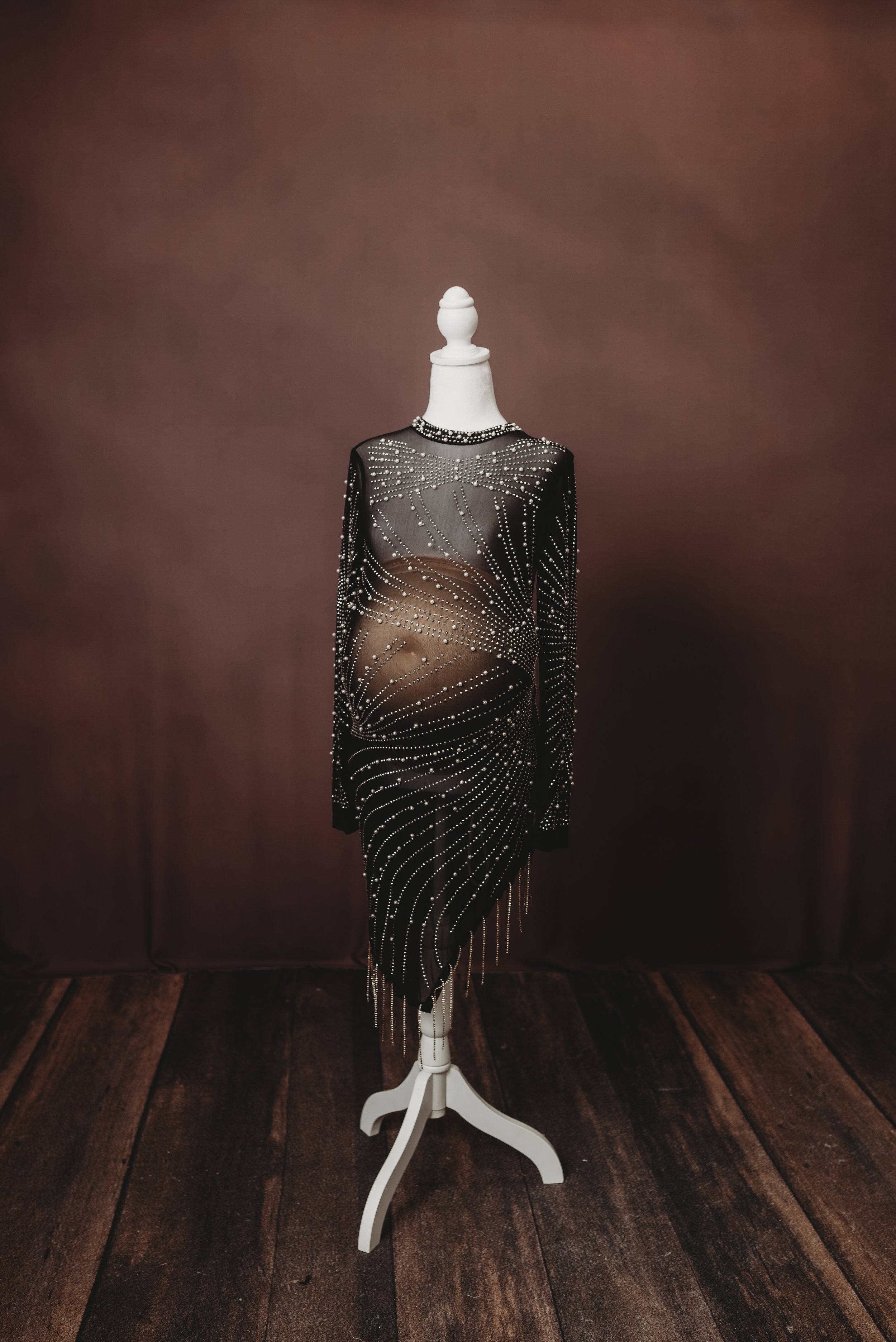 A mannequin dressed in a black, sheer, long-sleeved dress decorated with rhinestones and metallic beads, displayed on a white stand against a brown backdrop and wooden floor.