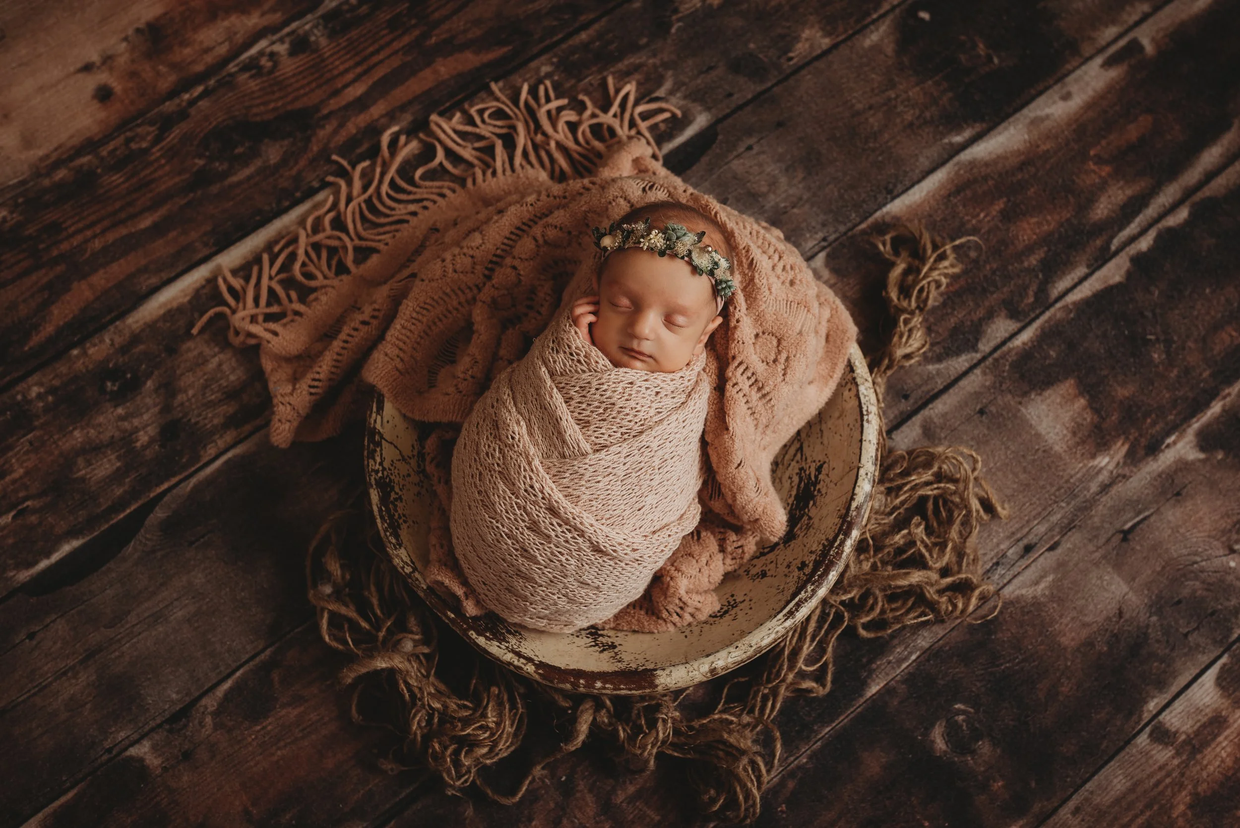 Newborn baby swaddled in a beige wrap, sleeping in a rustic bowl, with a floral headband, on a wooden floor with a fringed blanket.