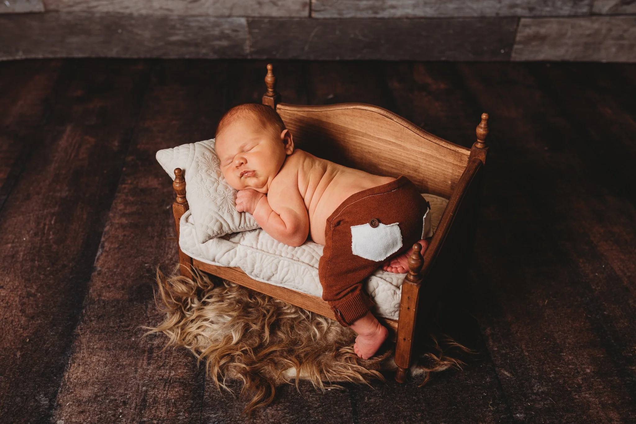 Newborn baby sleeping on a small wooden bed with a quilted pillow and brown pants, on a dark wooden floor.
