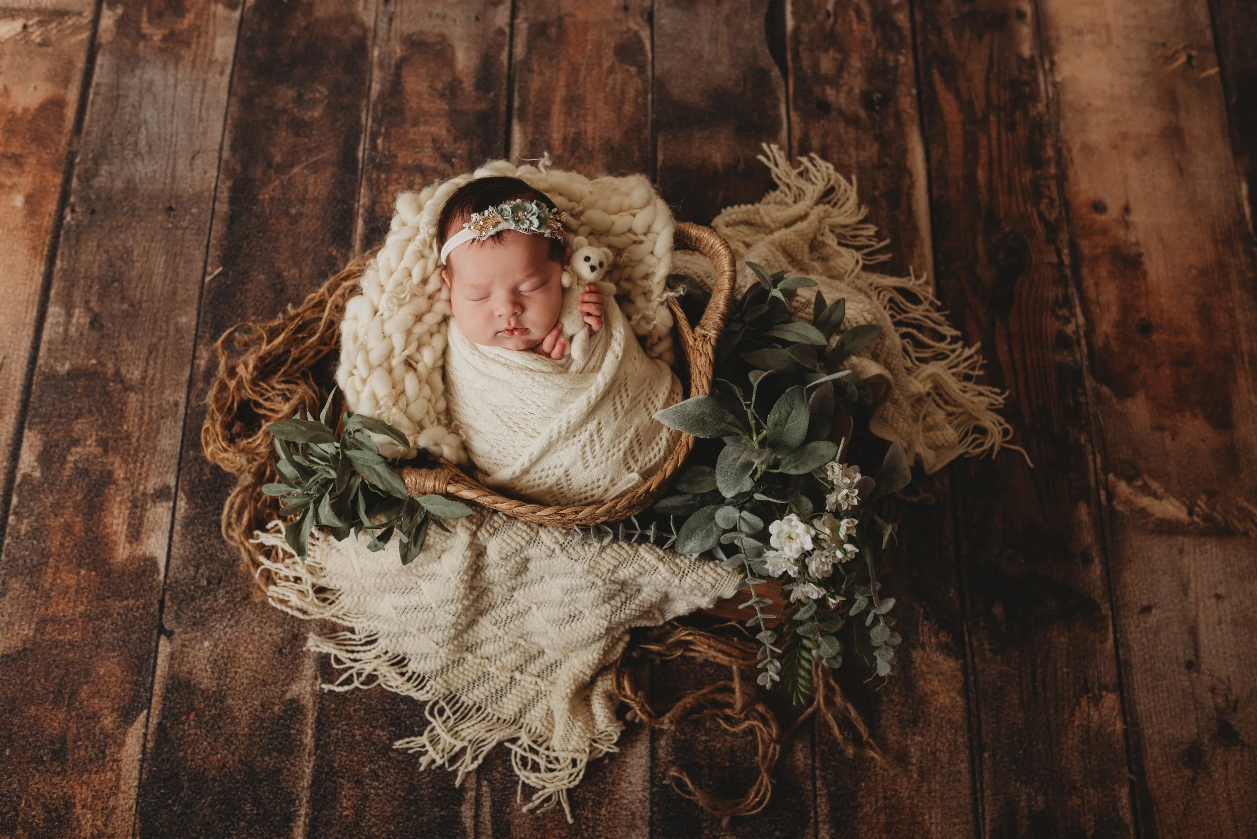 Sleeping newborn wrapped in a white blanket, lying in a wicker basket with green foliage and white flowers, on a wooden floor.