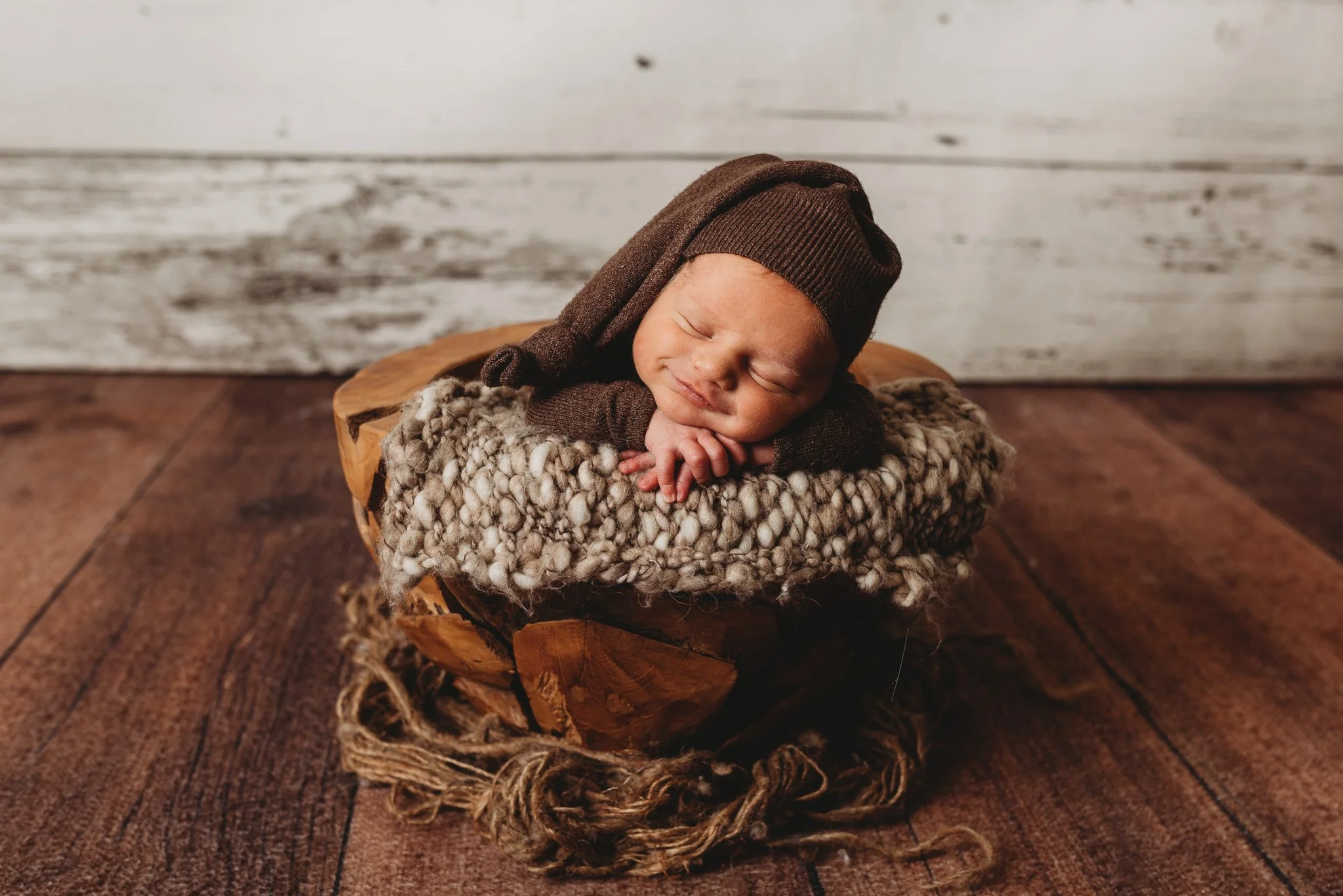 Newborn baby sleeping in a cozy knitted outfit on a wooden bowl with a textured blanket.
