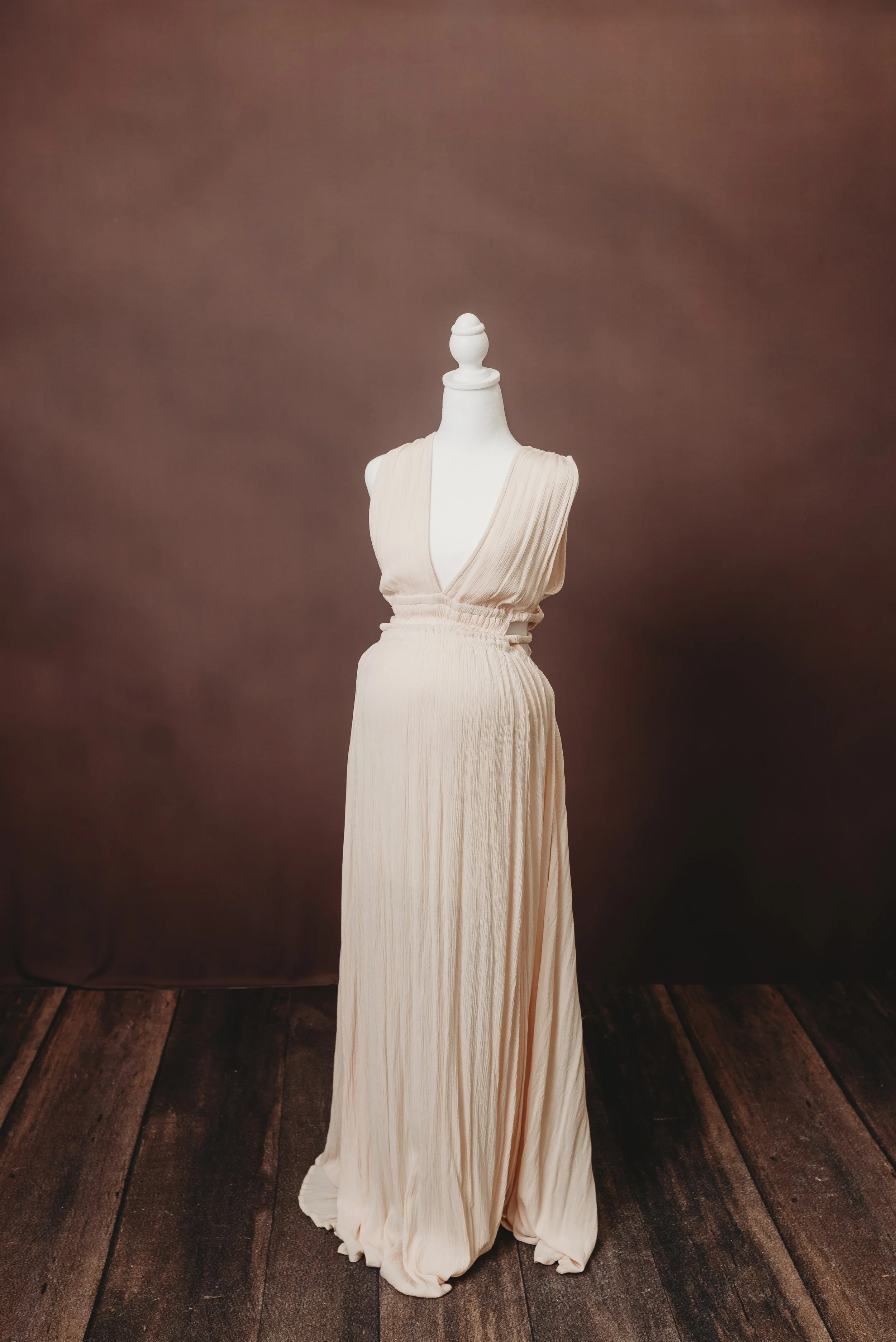A dress form mannequin wearing a long, flowing, off-white pleated dress, set against a plain brown backdrop with wooden flooring.