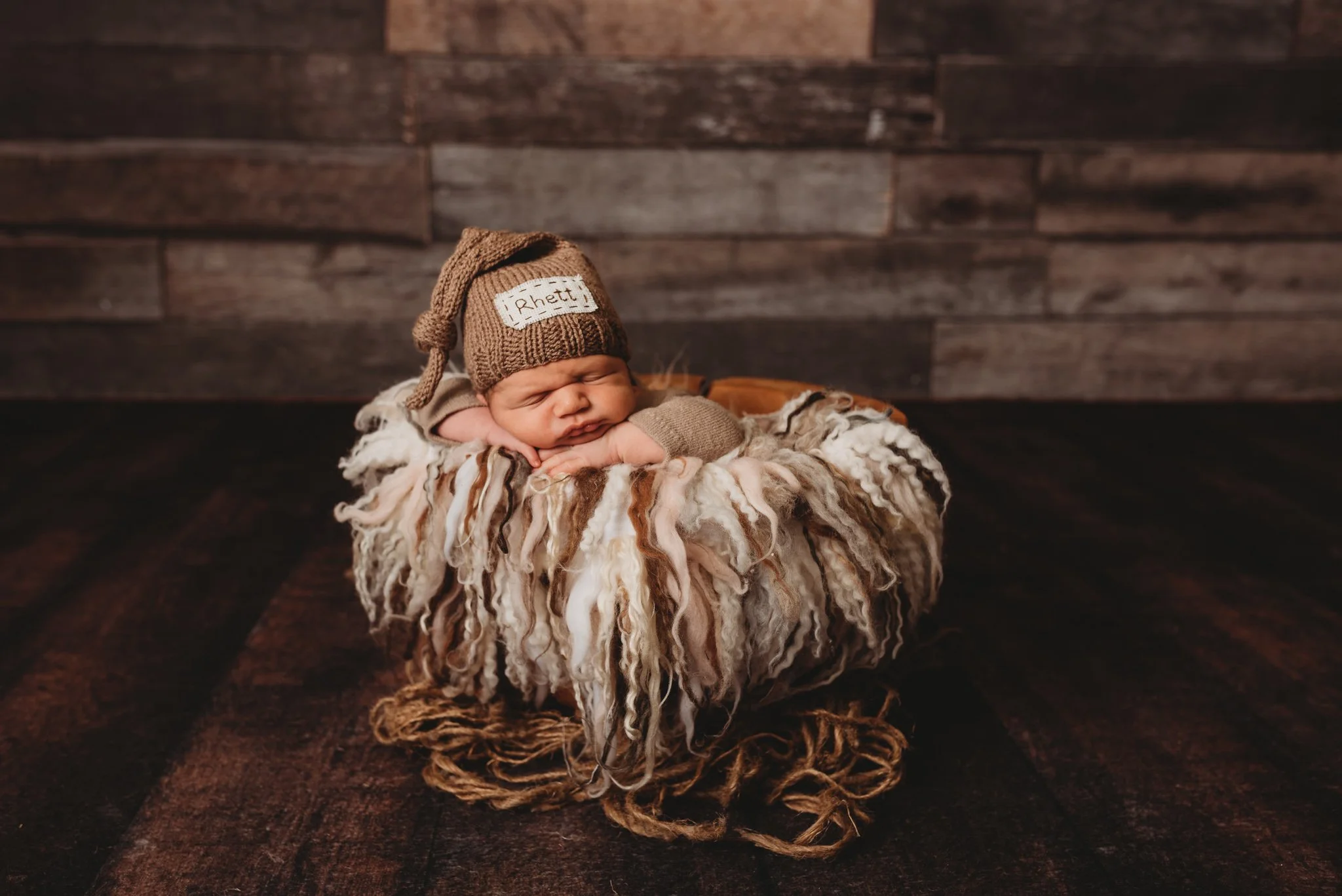 Newborn baby sleeping in basket wearing a knitted hat with "Rhett" label, wooden wall background.
