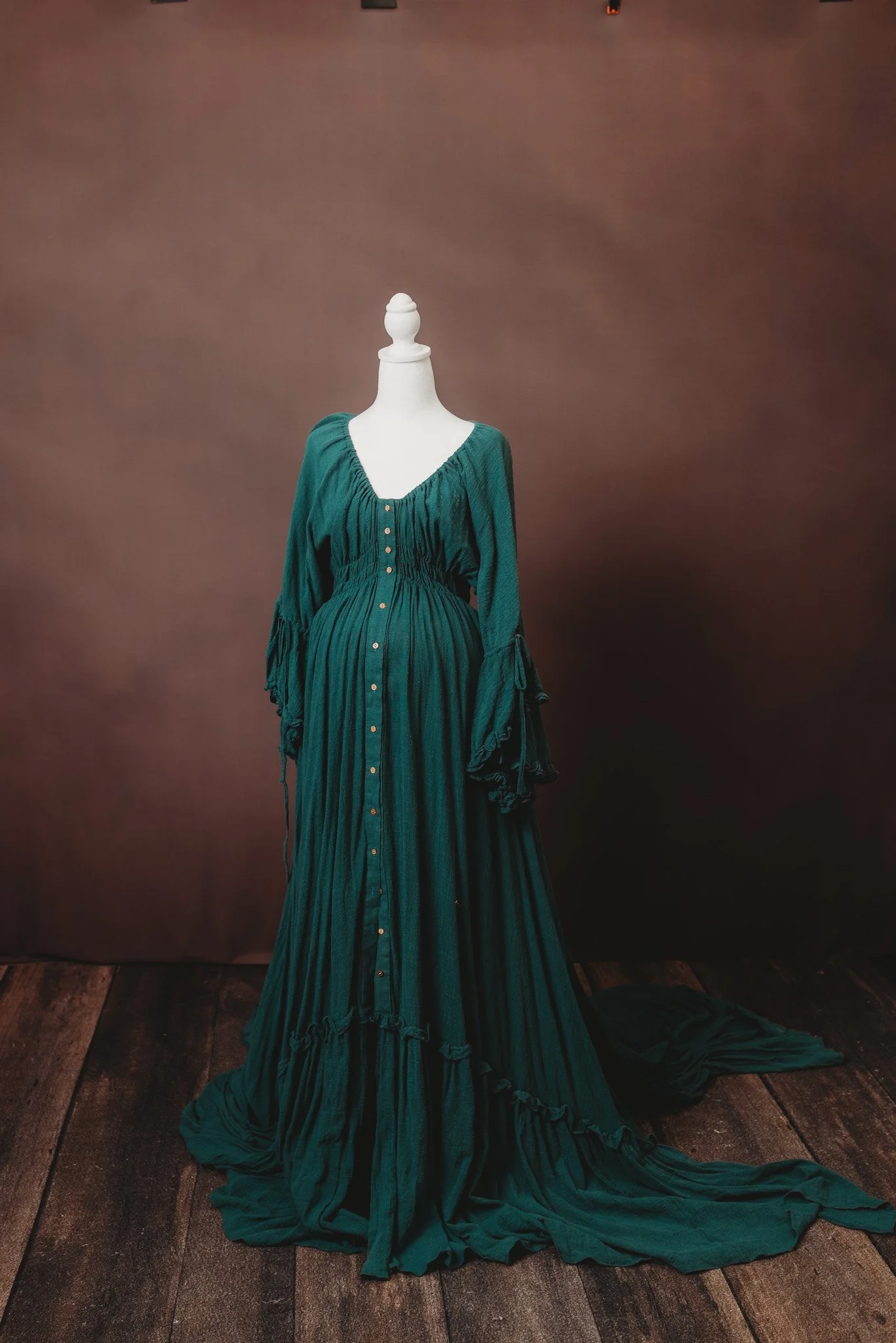 A mannequin wearing a long, dark green, pleated dress with button details, set against a plain brown background on a wooden floor.