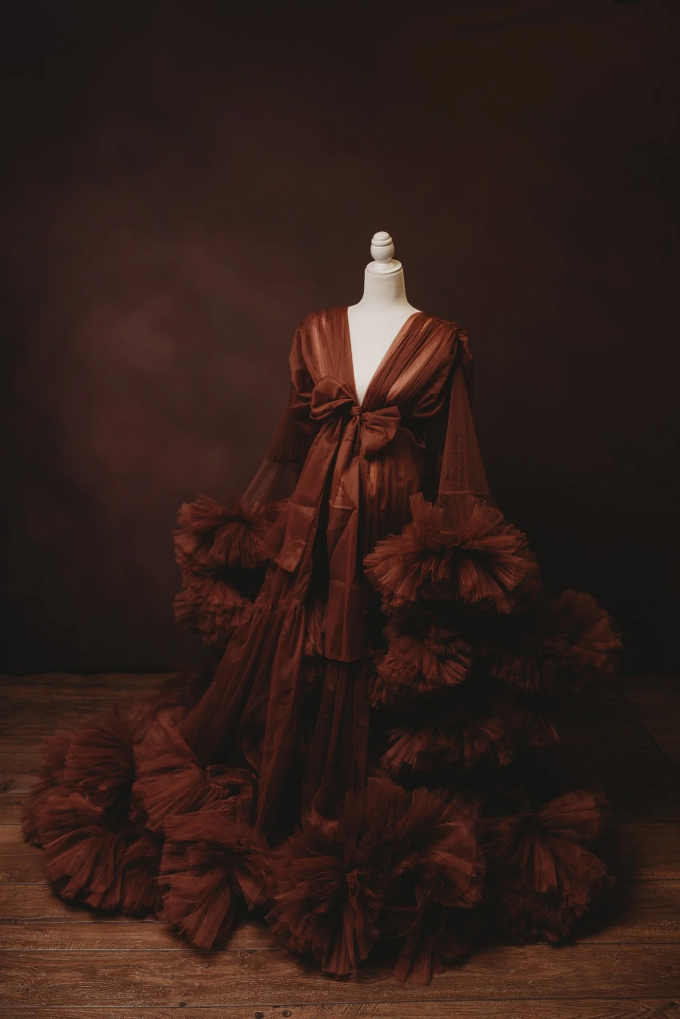 Elegant mannequin wearing a voluminous brown tulle gown with floral embellishments, displayed on a wooden floor against a dark backdrop.