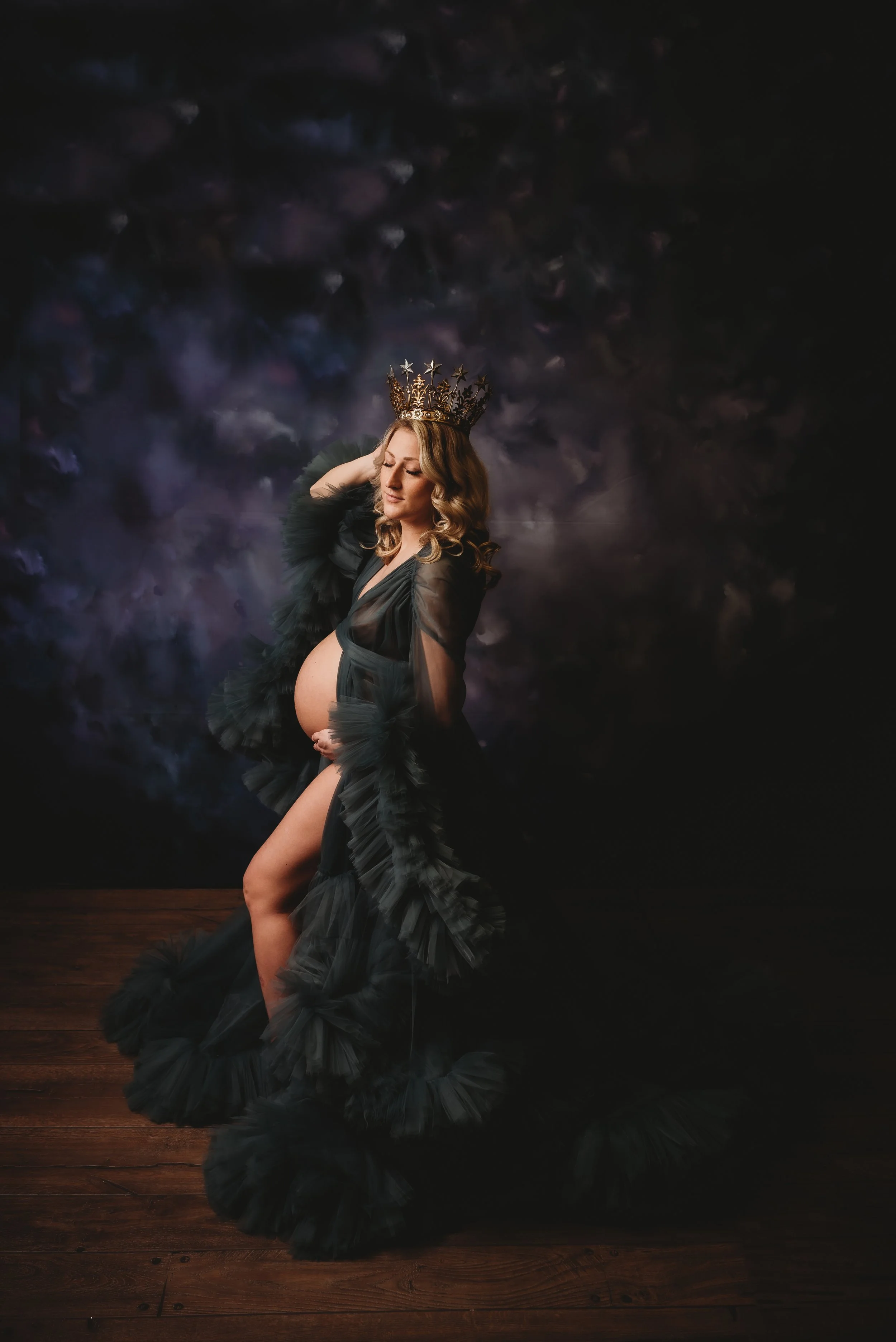 Pregnant woman in a flowy gown with a crown, standing on a wooden floor against a dark, textured background.