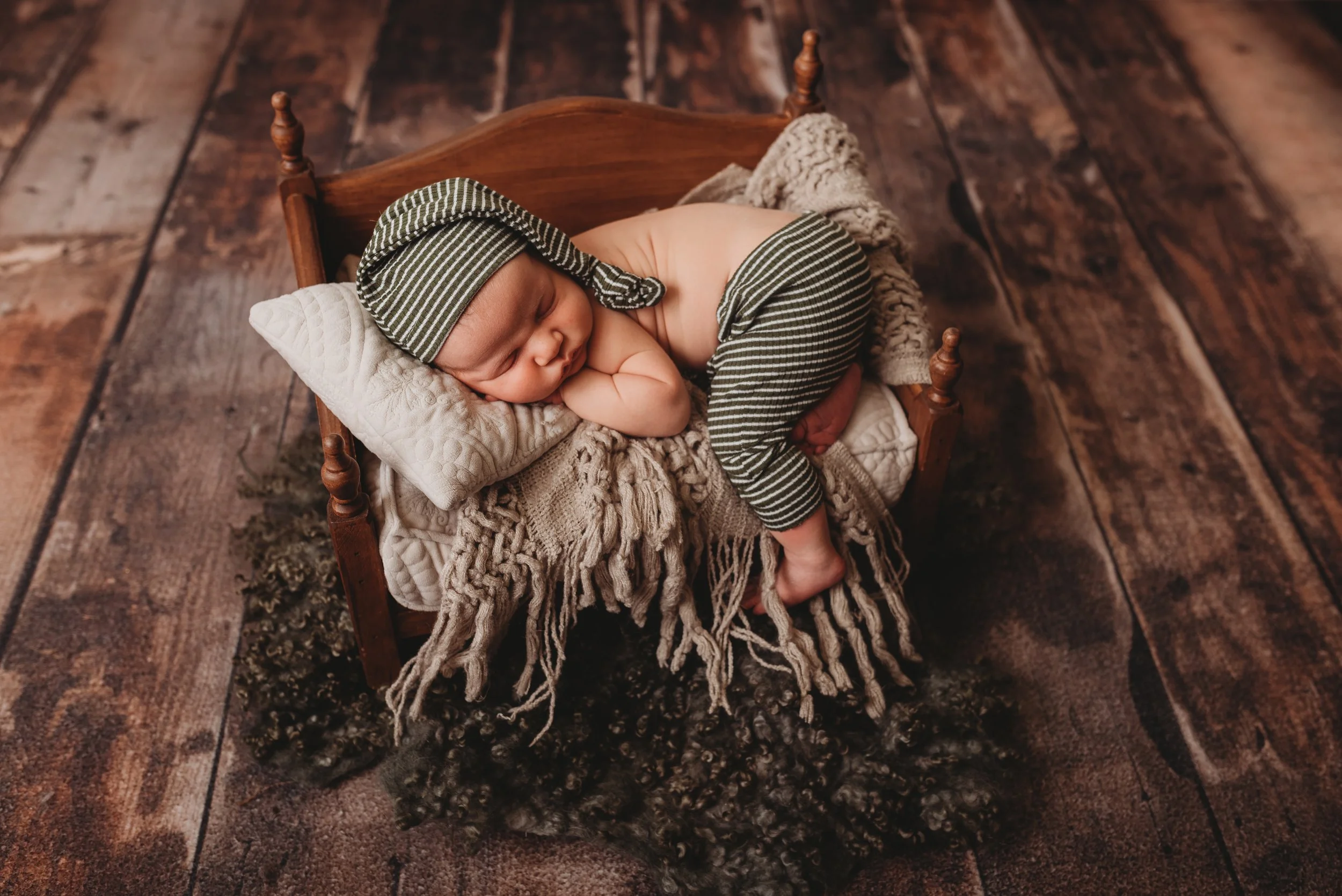 Newborn baby sleeping on small wooden bed, wearing striped hat and pants, cozy knit blanket.