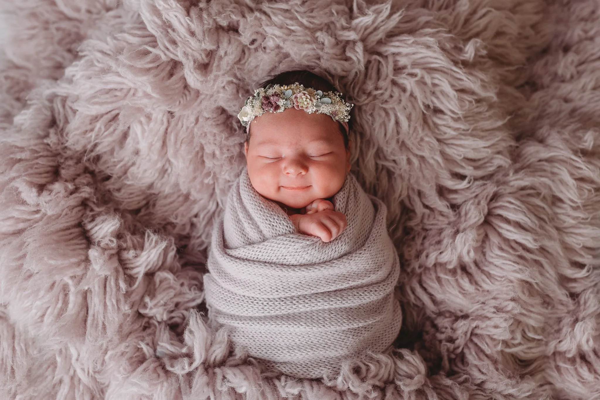 Newborn baby wrapped in a lavender knit blanket, wearing floral headband, lying on a fluffy surface.