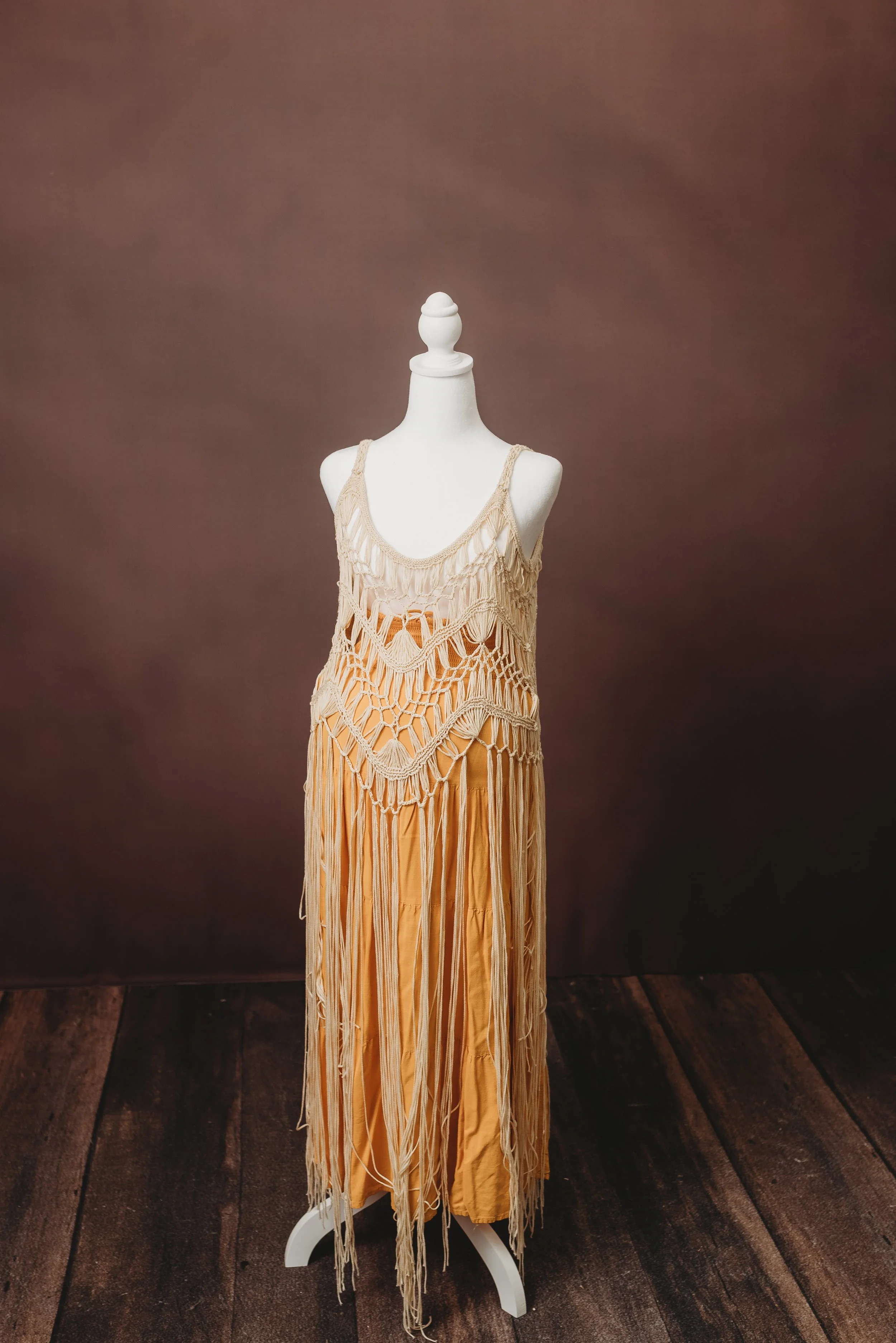 A mannequin dressed in a beige, handmade crochet dress with long strands of yarn hanging down, set against a brown wall.