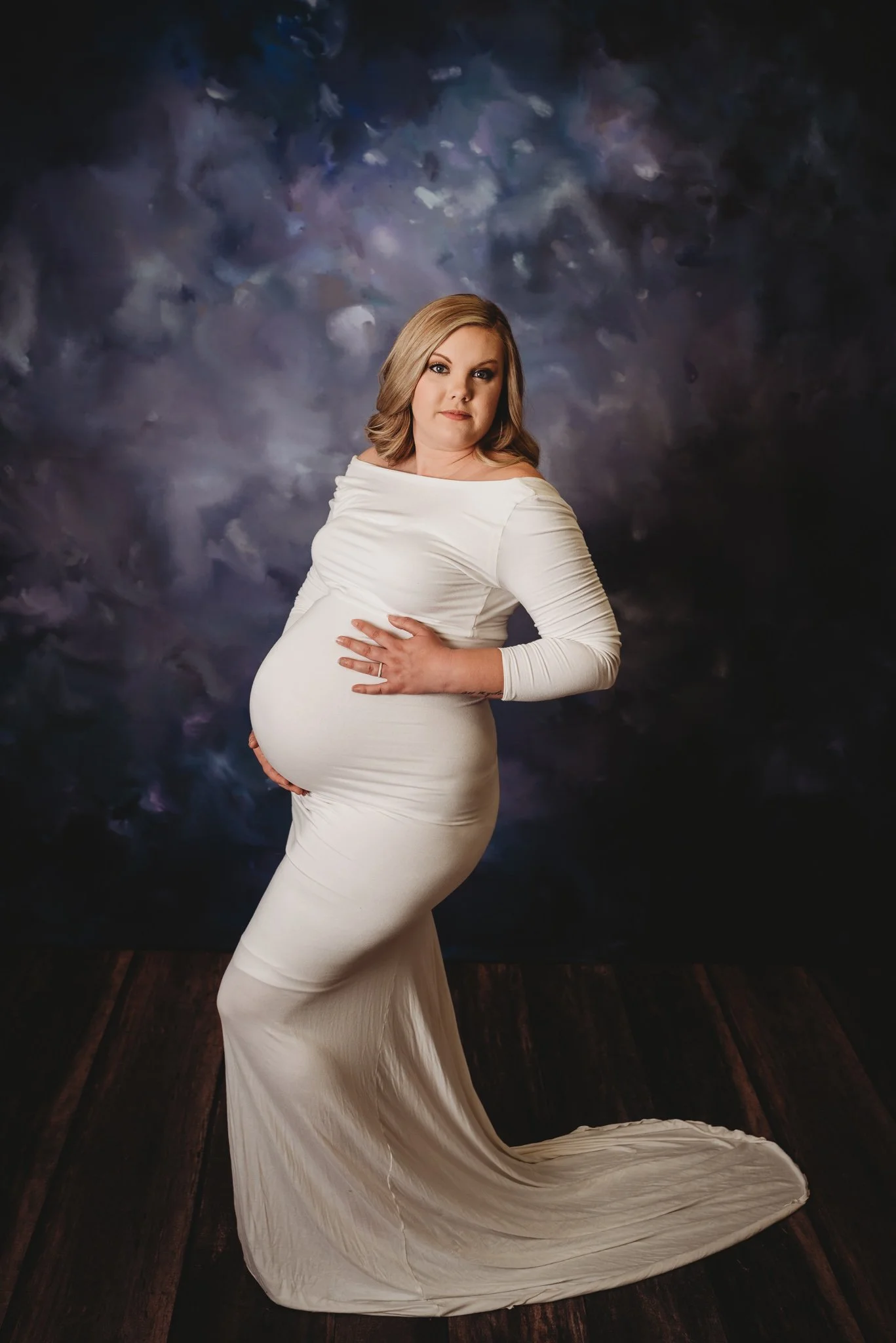 Pregnant woman in white dress posing against a dark textured background.