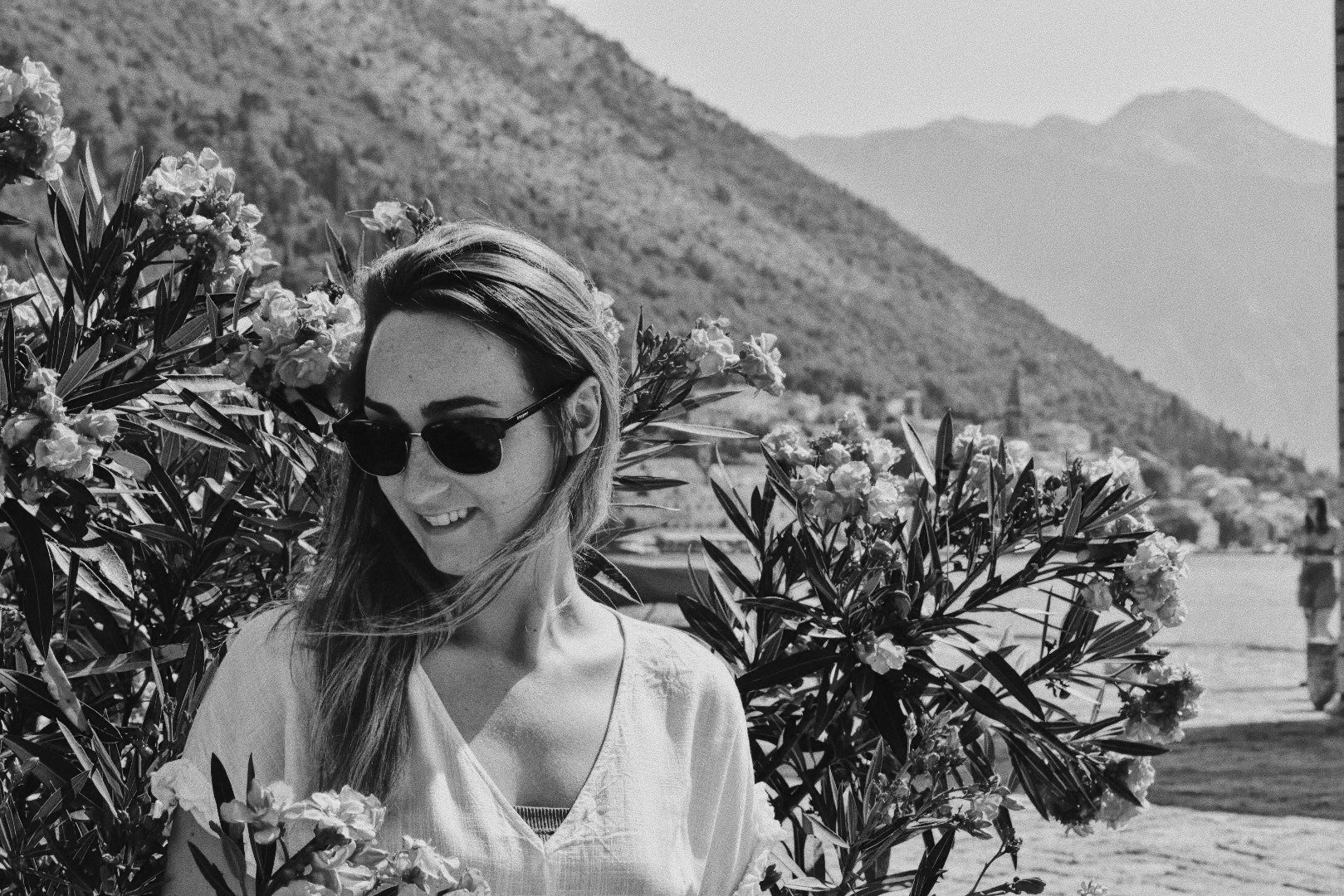 A woman wearing sunglasses and a light-colored top, smiling and looking down, standing among flowering bushes with mountains in the background.
