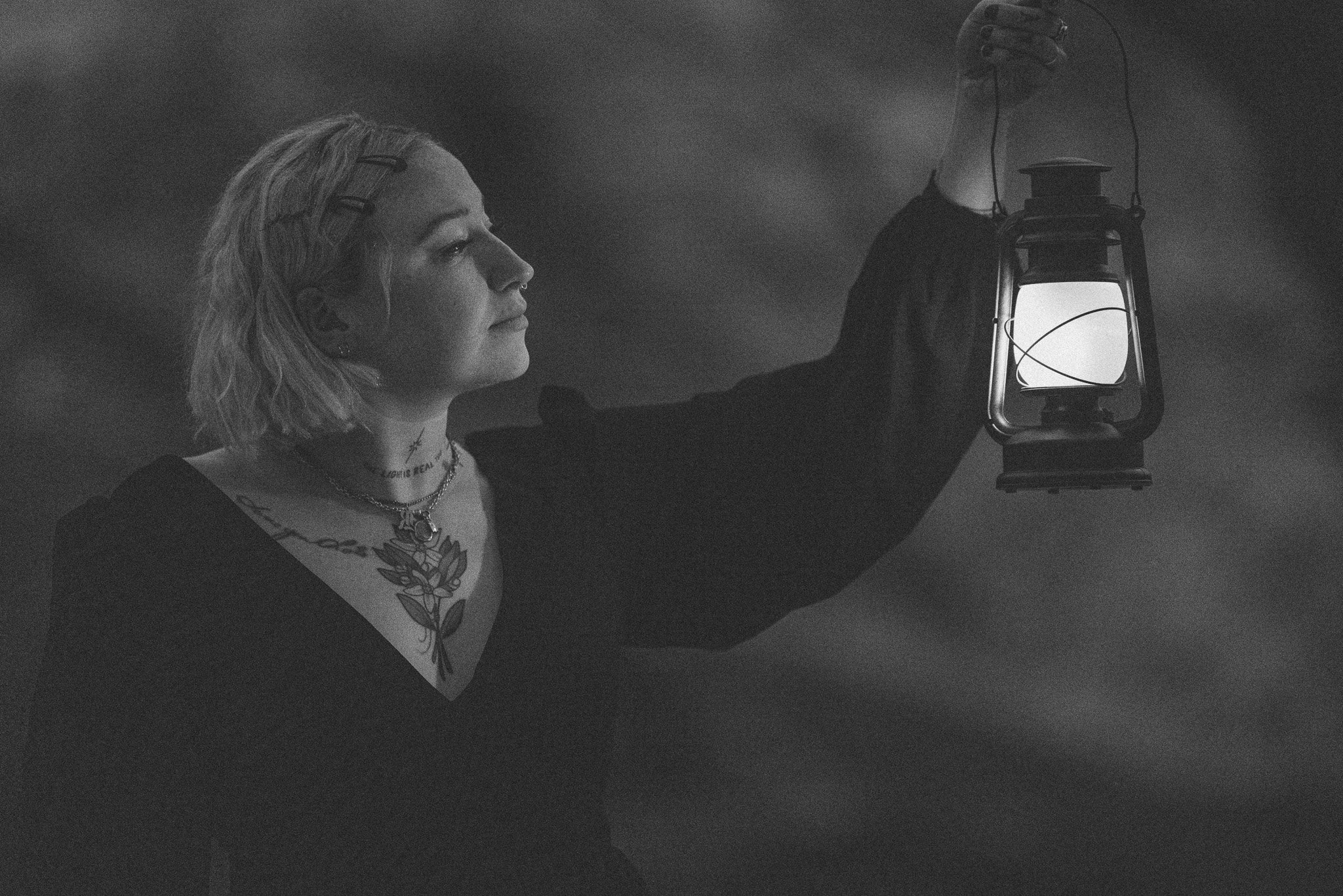 A woman with shoulder-length hair and tattoos on her chest holding an old-fashioned lantern in a dark room.