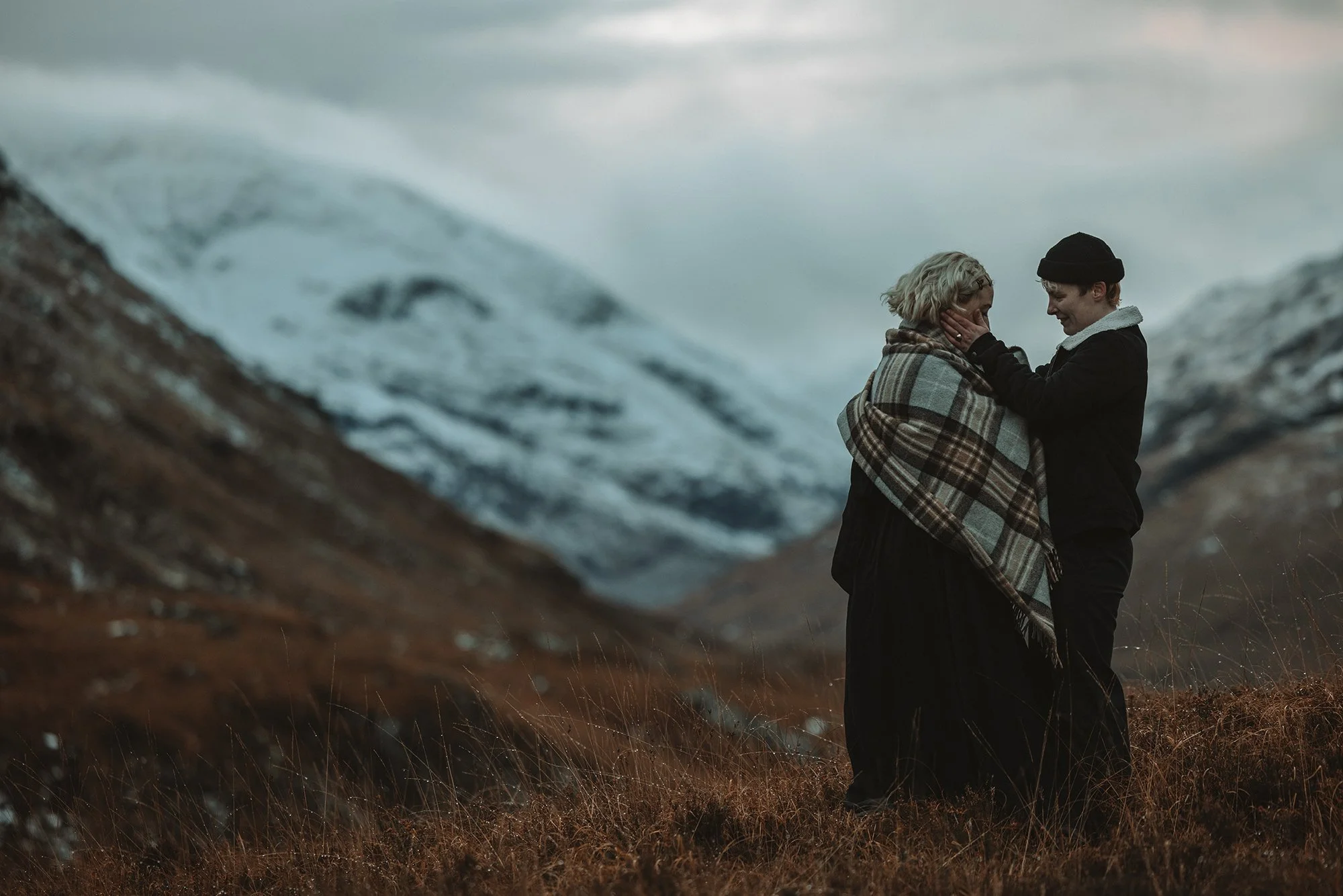 Two women standing close together outdoors in a mountainous landscape, with one woman gently touching the other's face. They are dressed warmly, one with a plaid shawl and the other in a black coat and beanie, amidst brown grass and overcast skies.