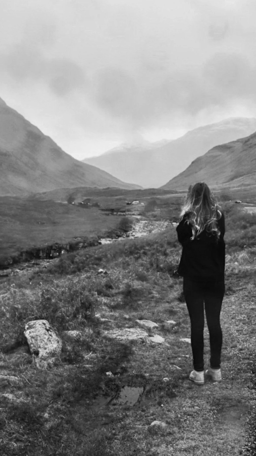 A woman with long curly hair stands outdoors on a trail in a mountainous landscape, facing away from the camera, in black and white.