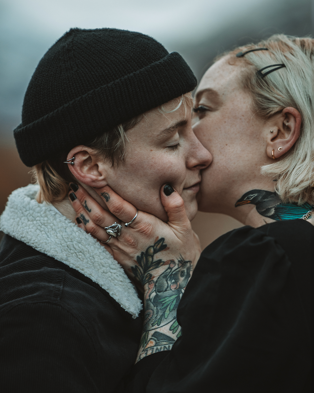 Two people share an intimate moment, one with tattoos and piercings, holding the other’s face while they lean in for a kiss. The person with tattoos has short, light hair and wears a black beanie and a black jacket with a shear-lined collar. The other person has blonde hair with glasses on top and has visible freckles. The background is blurred and cloudy.
