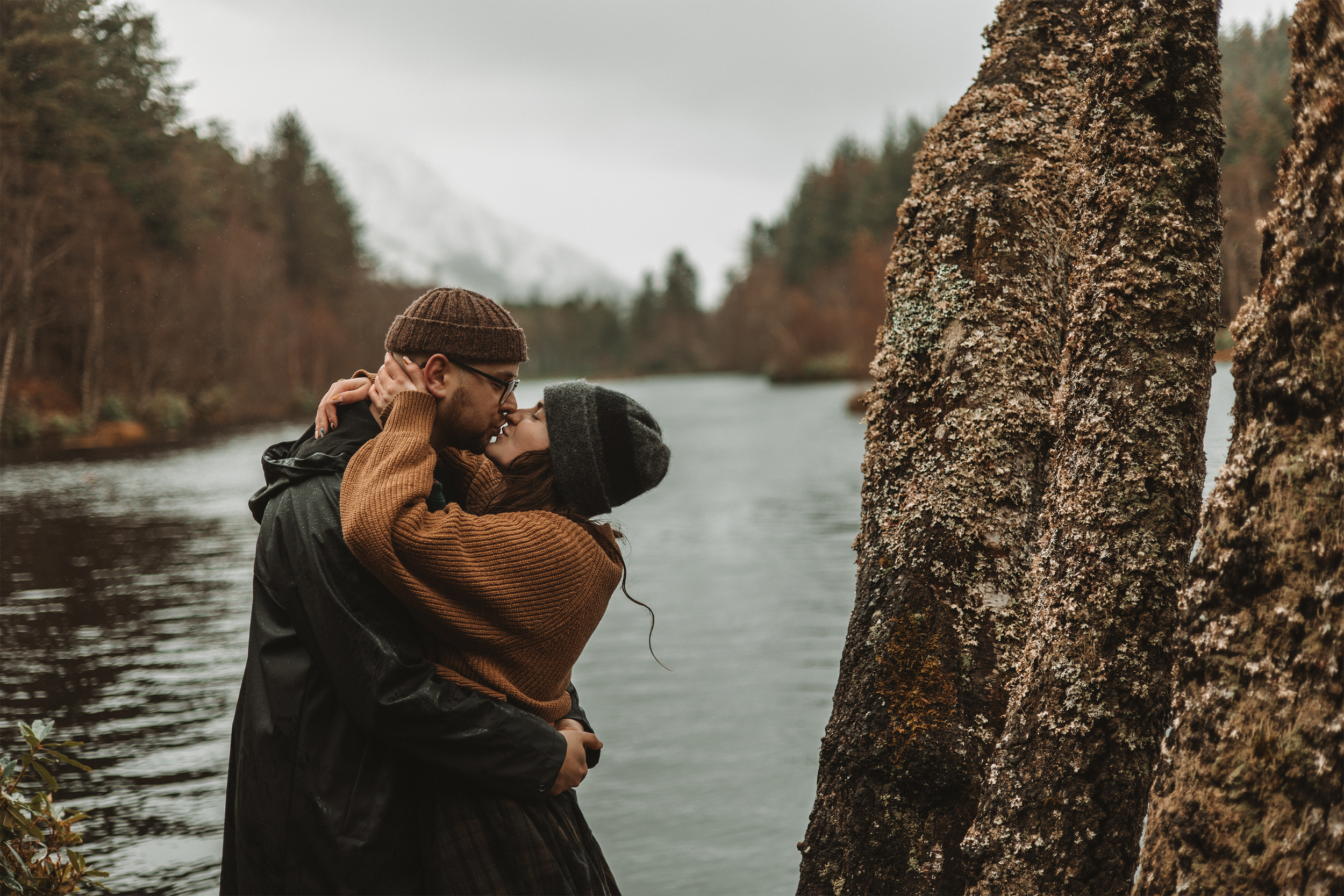 A couple stands close by a tree near a river, sharing an intimate moment, with a natural landscape of trees and mountains in the background on a cloudy day.
