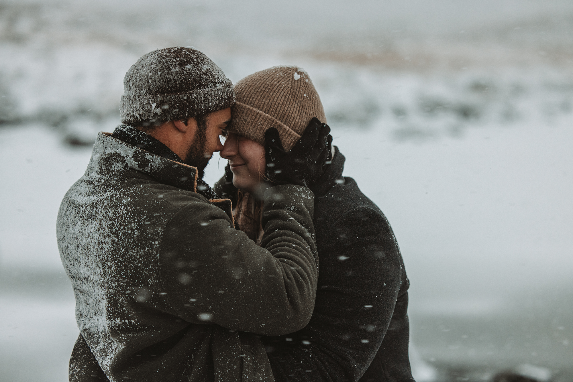 A couple in warm clothing share a tender moment on a snowy day, with their foreheads touching and eyes closed.