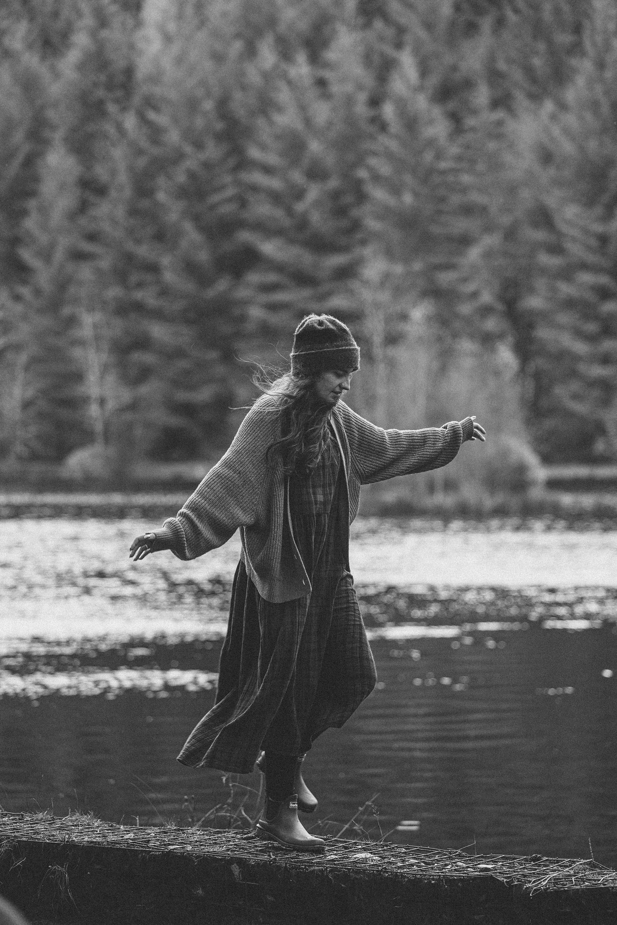 A woman walking on a narrow wooden bridge by a river in a forest, wearing a hat, sweater, and skirt.