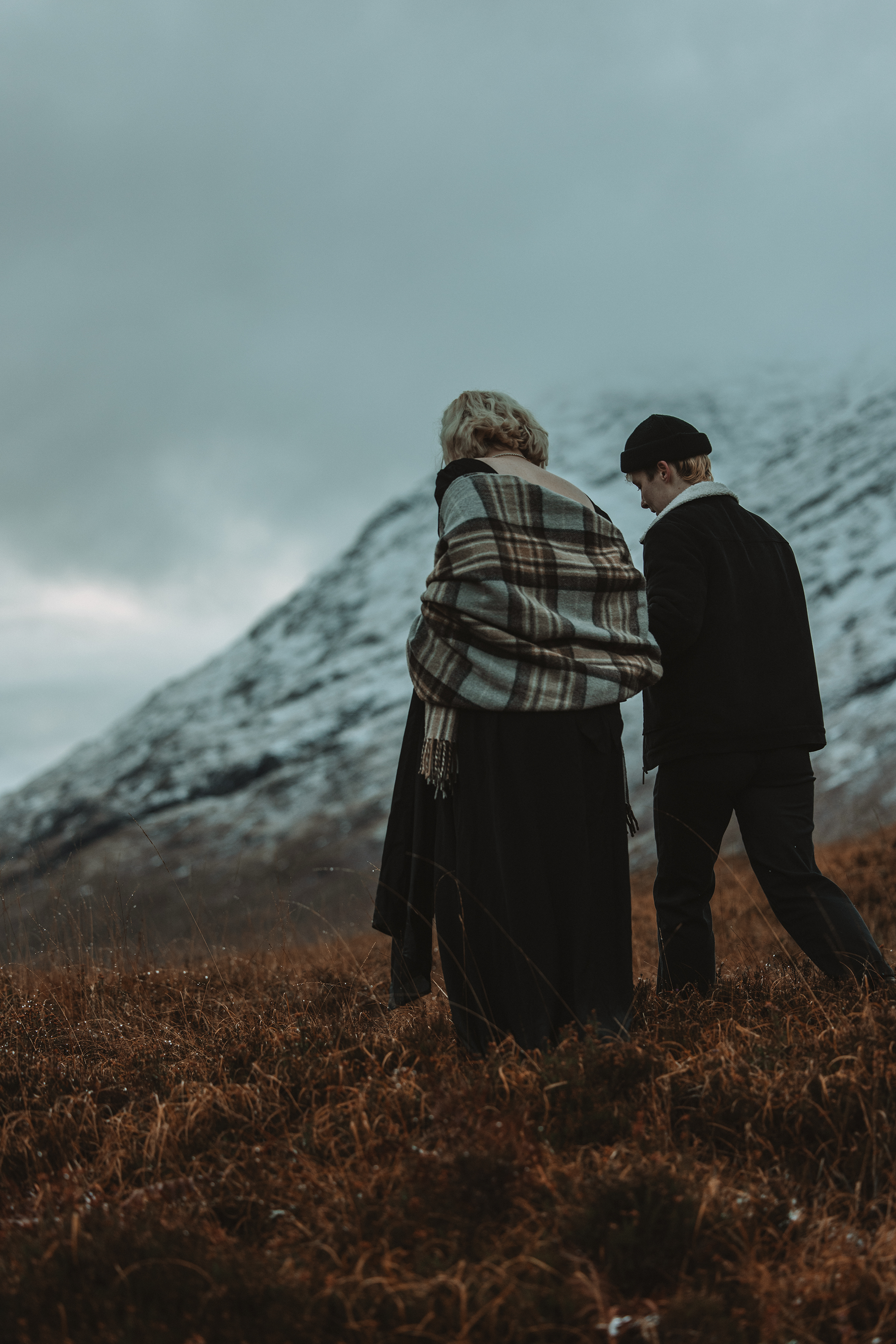 Two people standing in a grassy field with mountainous terrain in the background, overcast sky.