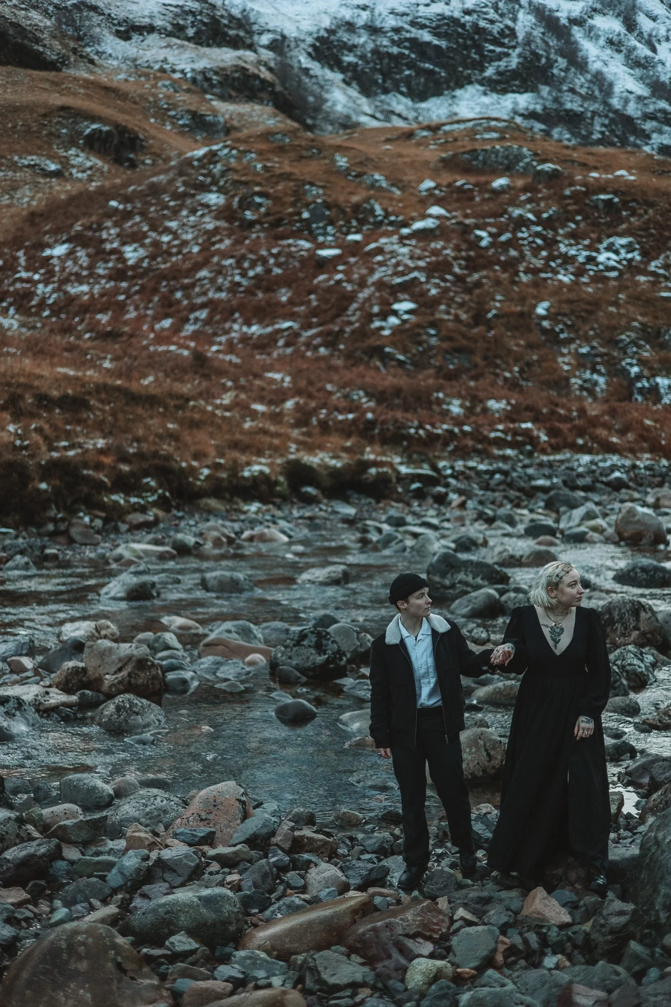 Two women walk along a rocky riverbank surrounded by mountains with snow and brown vegetation.