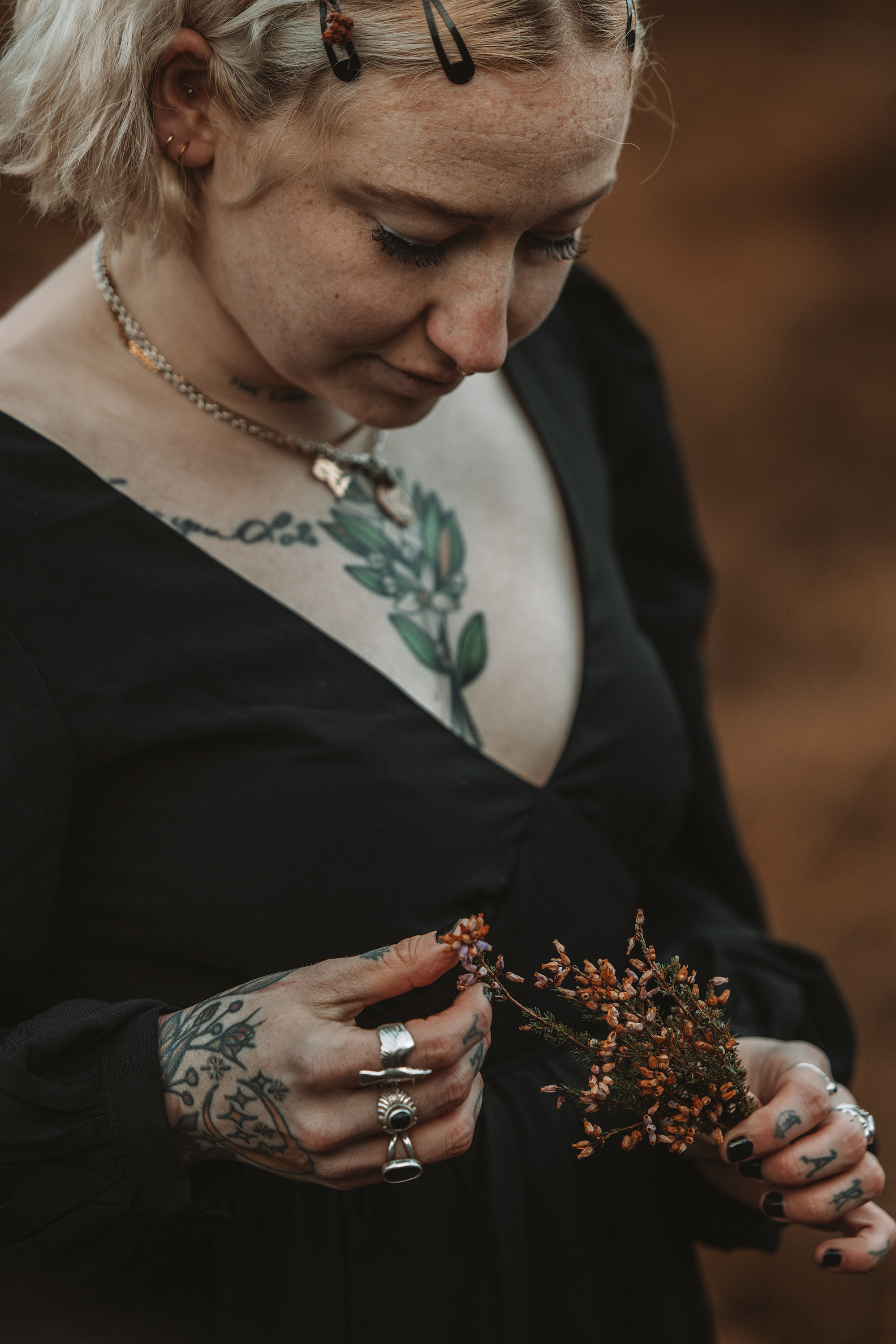 A person with short blond hair, tattoos, and multiple rings on their fingers holds a small bouquet of dried flowers. They are wearing a black top and have several necklaces, including one with a quote and another with a pendant. Their face is angled 