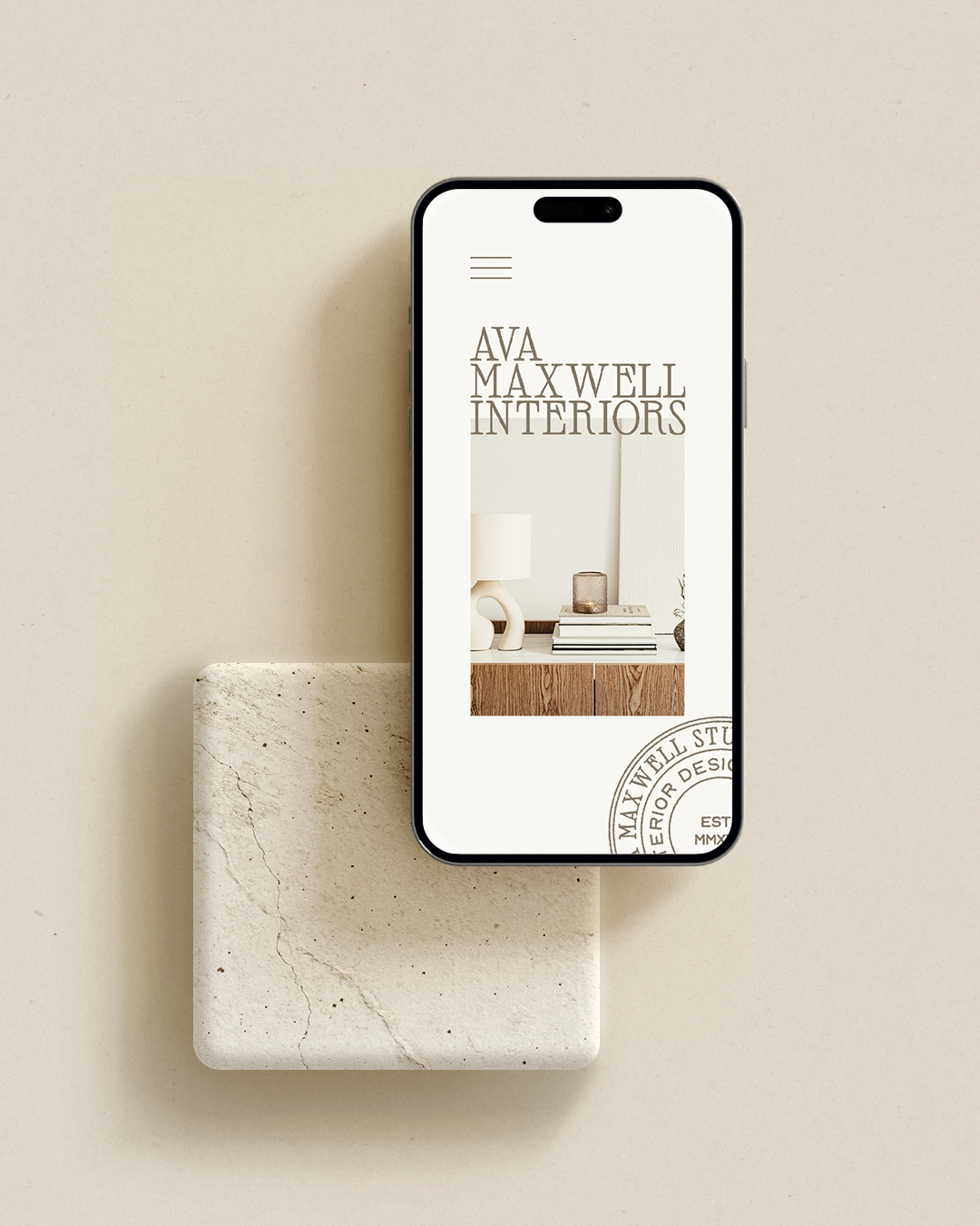 Smartphone displaying 'AVAMAXWELL INTERIORS' website with a minimalist interior design image and logo, placed on a textured beige stone slab against a beige background.