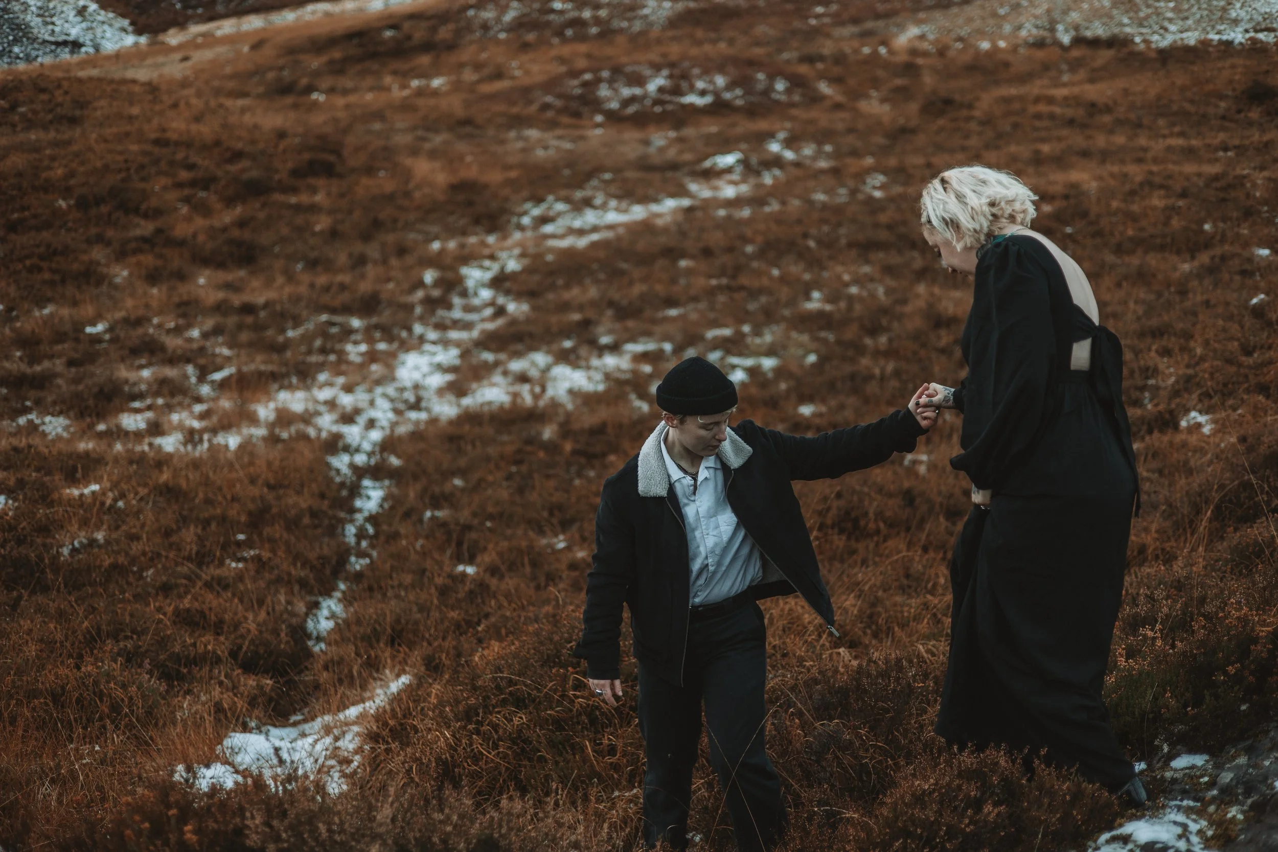 Two young people outside on a hillside with patches of snow, one with blonde curly hair wearing dark clothing and the other with dark hair and a hat, holding hands.