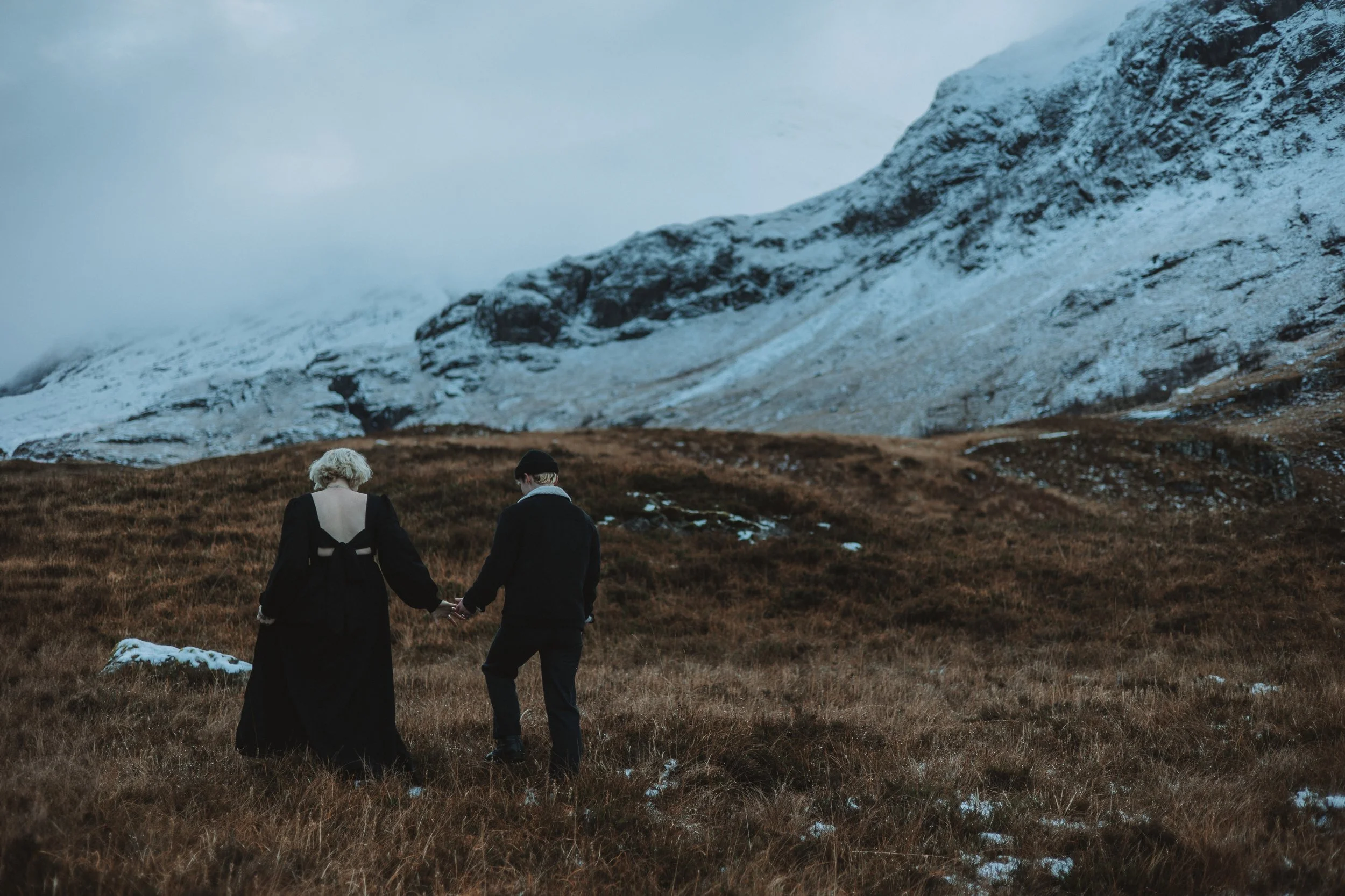 A couple holding hands walks through a grassy field with patches of snow, with snow-capped mountains and cloudy sky in the background.