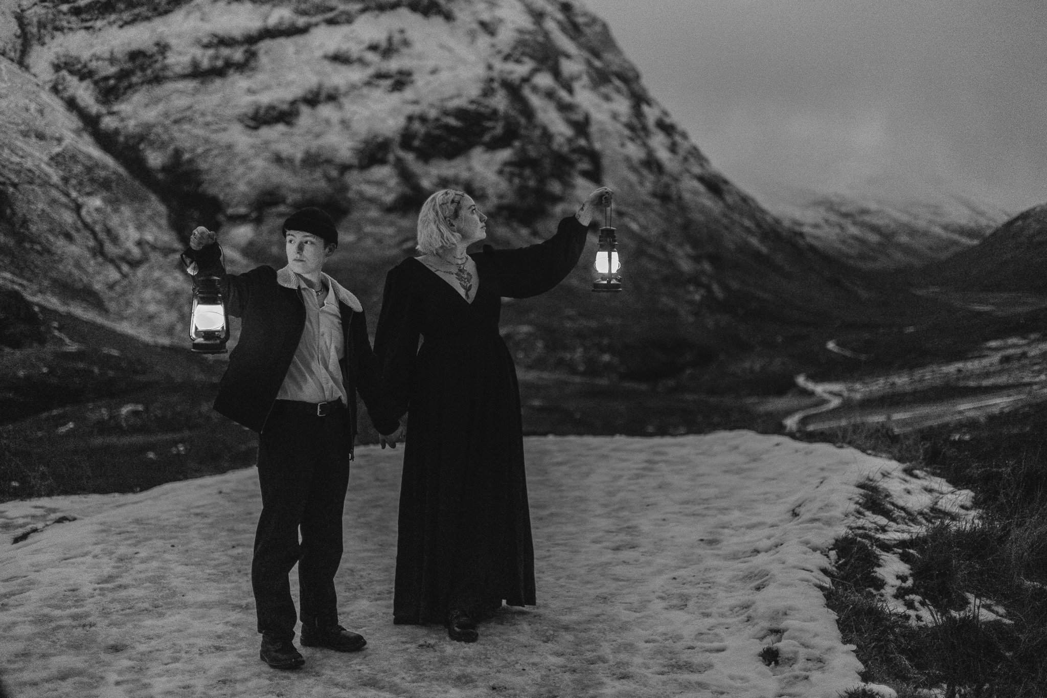 Two women stand outdoors in a snowy landscape with mountains in the background. They hold lanterns and look ahead, one is dressed in a long black dress and the other in a jacket and shirt.
