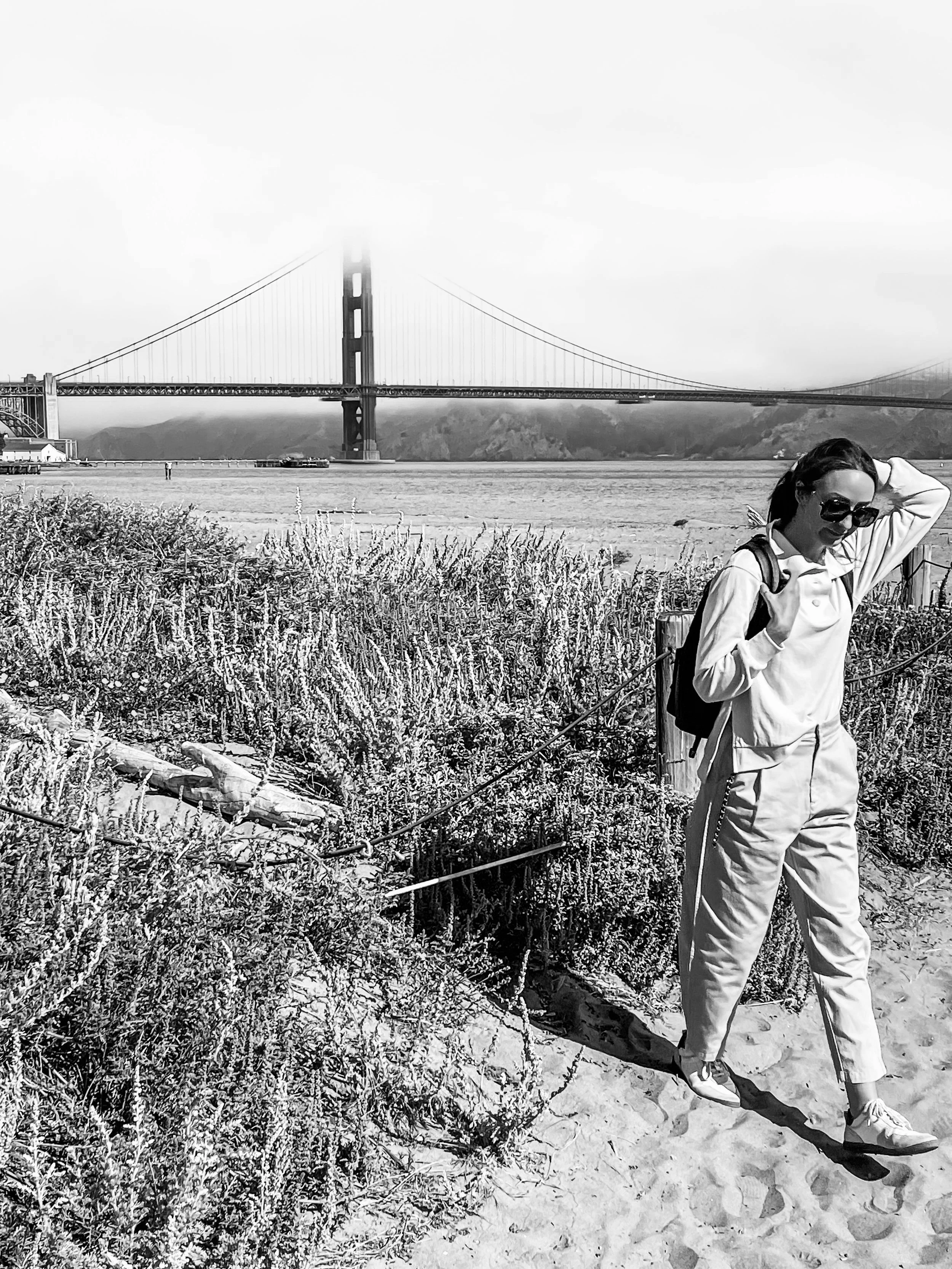 Woman standing on sandy beach by water with Golden Gate Bridge in background, wearing sunglasses and a backpack.