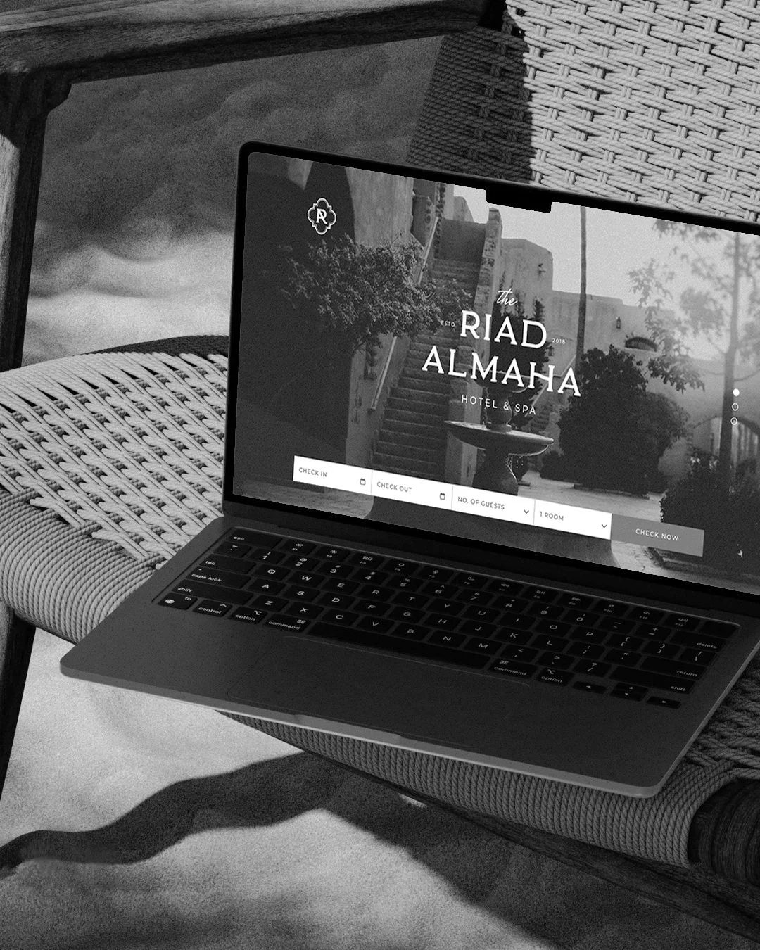 A laptop resting on a woven outdoor chair shows the hotel and spa website, 'The Riard Almaha,' with options for check-in, check-out, number of guests, and room selection, set outdoors with trees and a stone wall in the background.