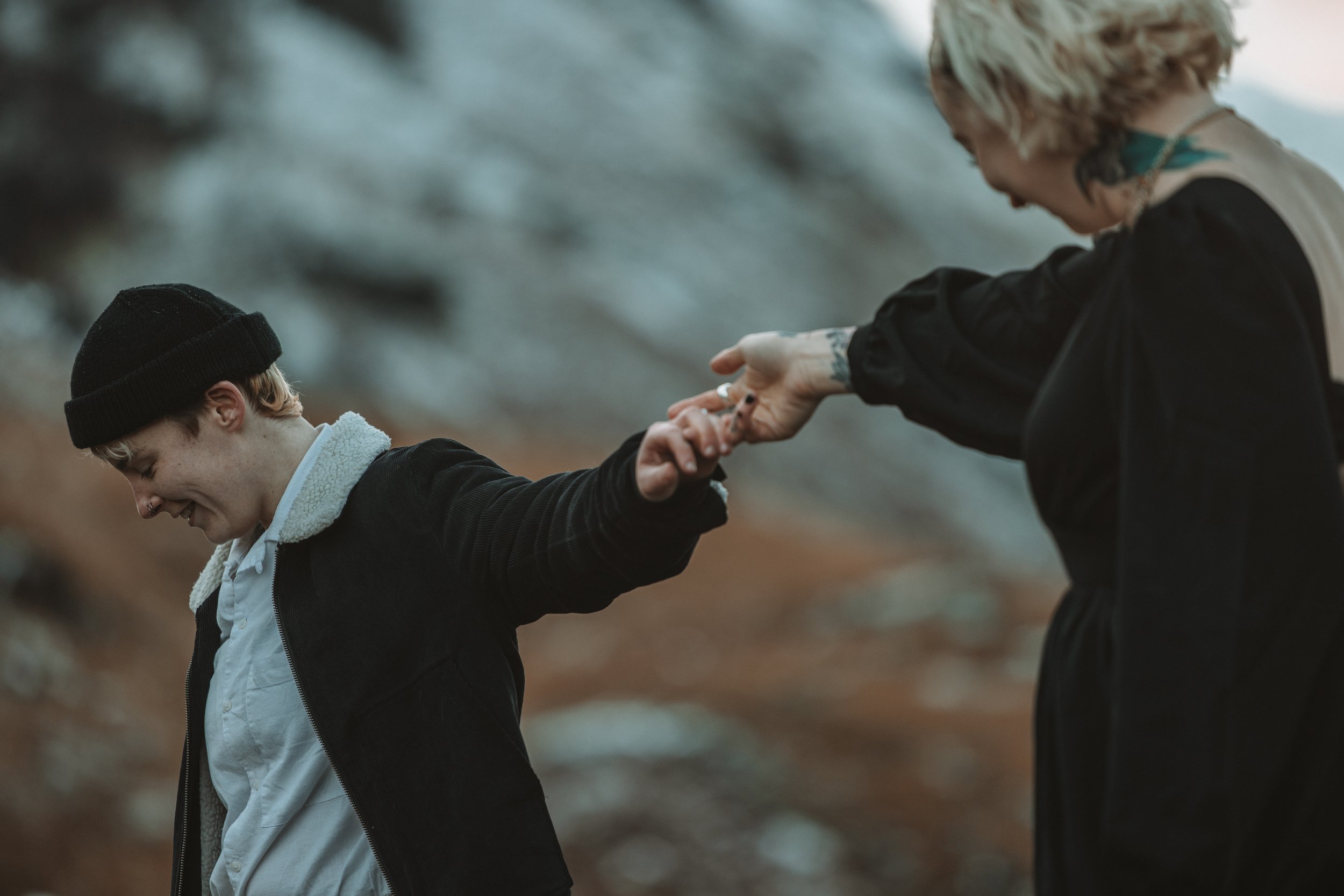 A person with tattoos and curly blond hair helping a younger person with light brown hair, wearing a black beanie, in an outdoor setting with blurred trees in the background.