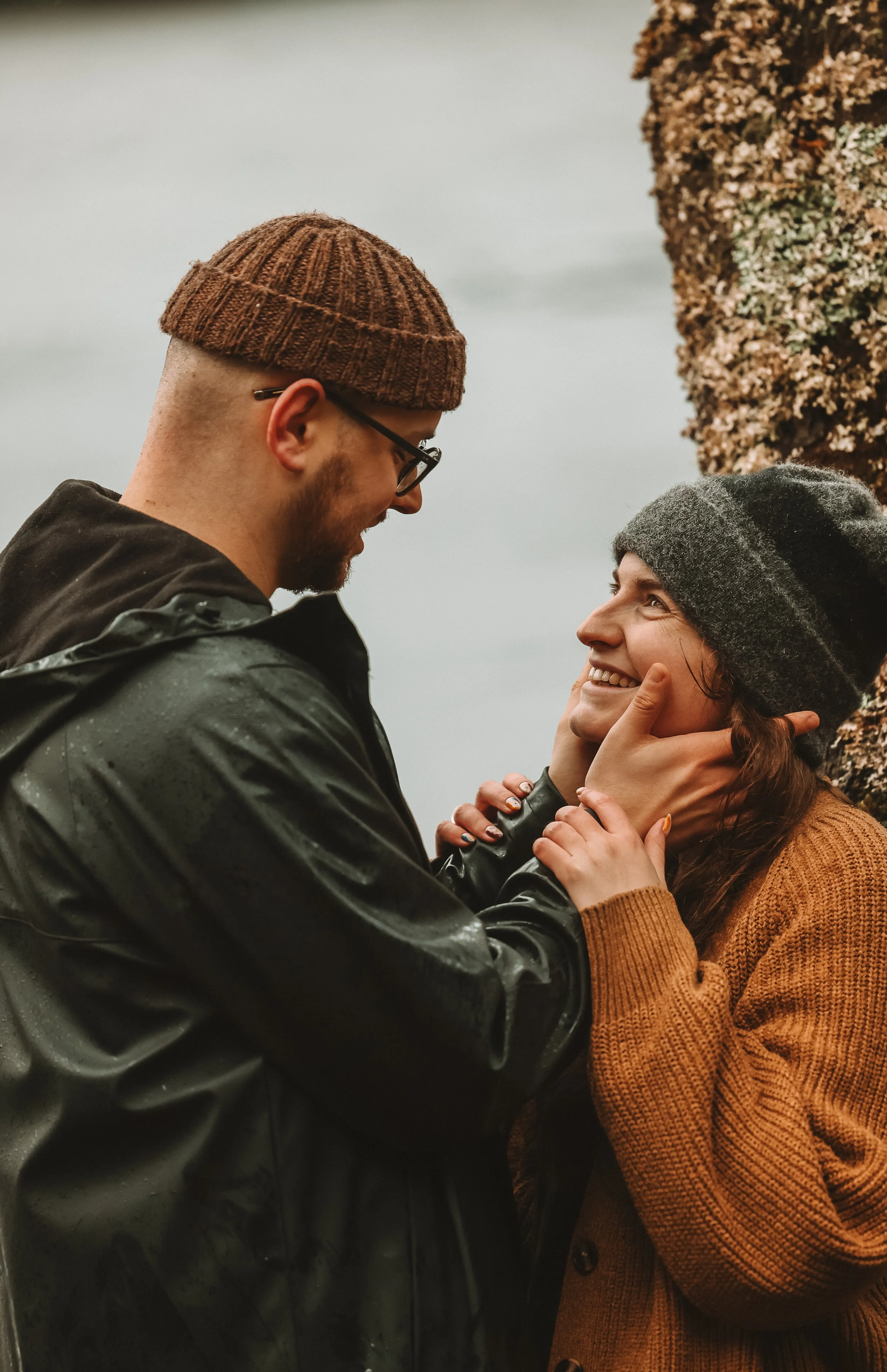 A man and woman facing each other outdoors during rainy weather. The man wears glasses, a brown knit beanie, and a black raincoat. The woman wears a gray knit hat and a brown sweater. They are smiling and touching each other's faces gently, with a tr