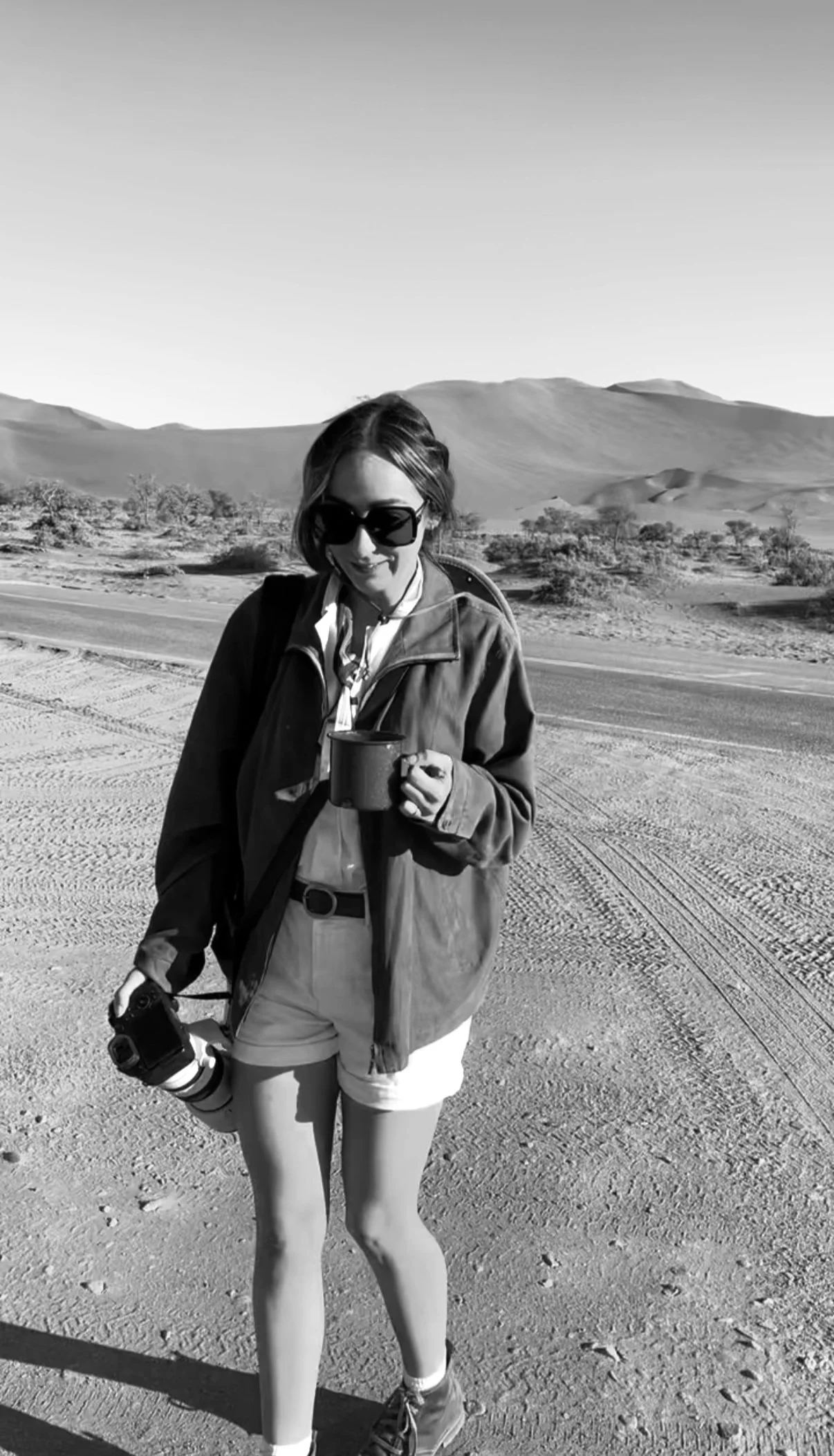 A woman standing in a desert landscape with mountains in the background, holding a camera and a mug, wearing sunglasses, a jacket, shorts, and hiking boots.