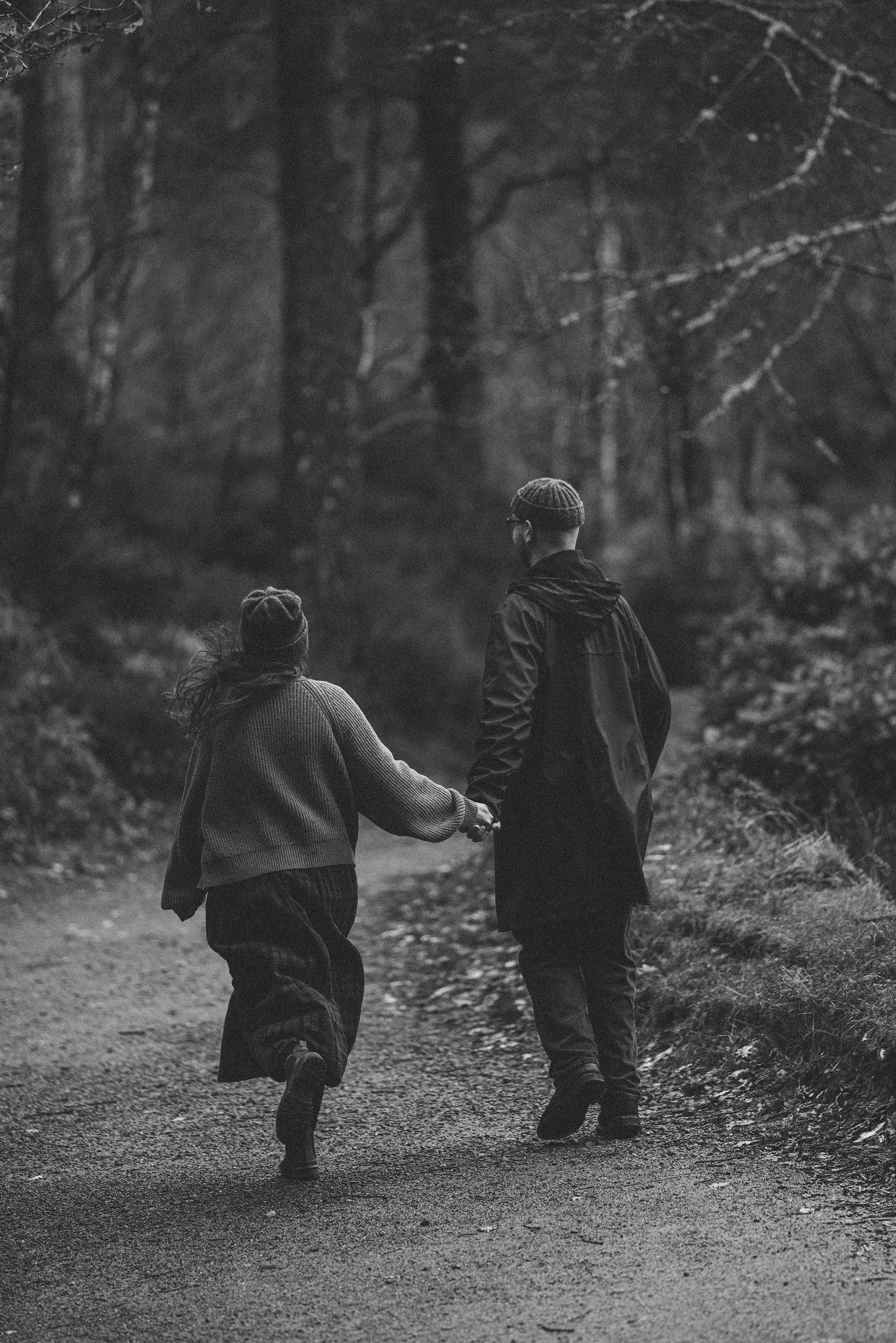 A black and white photo of a man and woman walking hand in hand on a forest trail. The man is wearing a beanie and glasses, and the woman has long hair and a hat. Trees surround the path, creating a peaceful, natural setting.